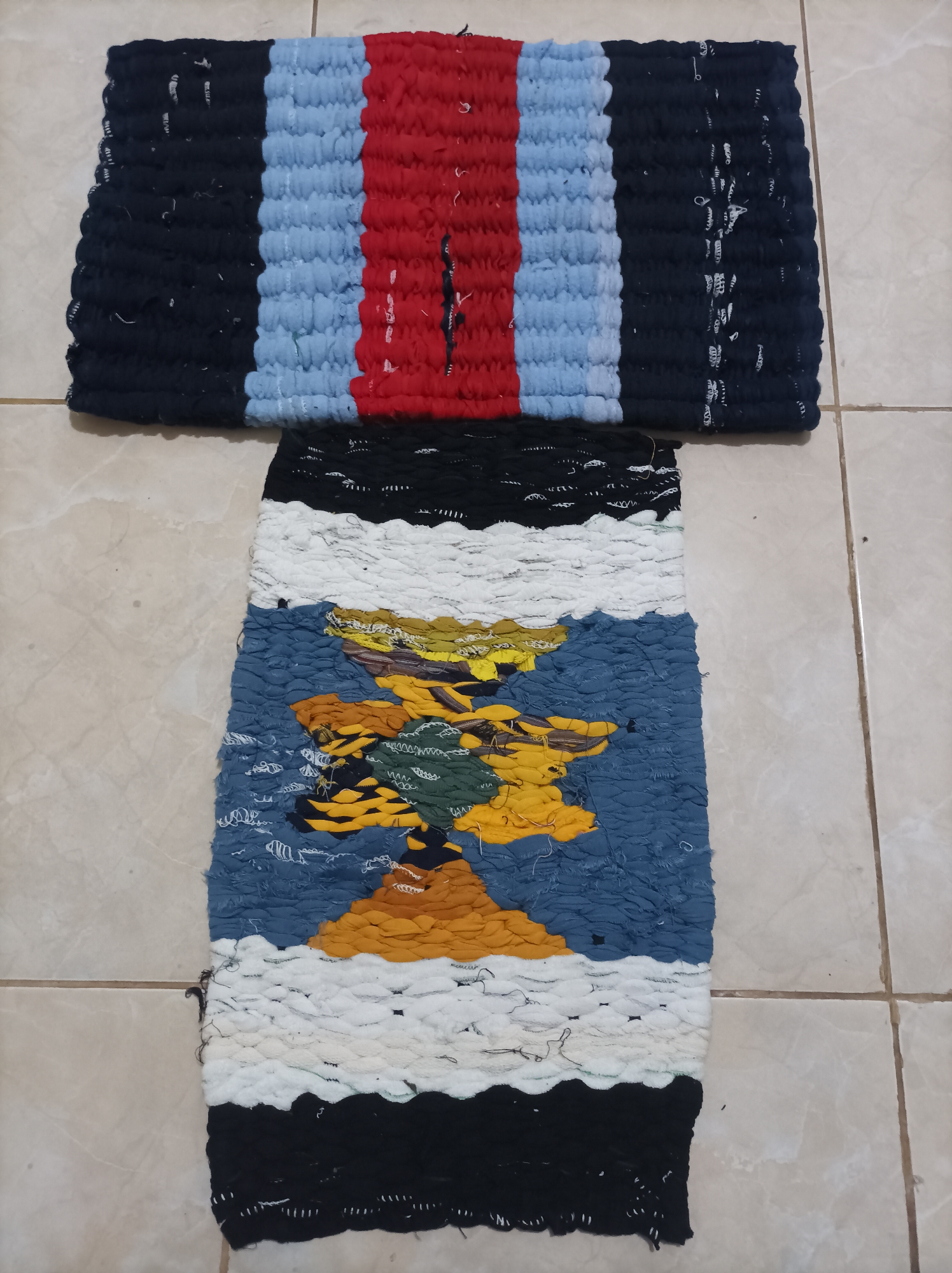 The image displays two handmade rugs laid out on a tiled floor. The upper rug is rectangular and features horizontal stripes of black, light blue, and red. The red stripe has a vertical black element incorporated into it. The lower rug is also rectangular but appears to be made with a more intricate weaving technique, showcasing a colorful abstract pattern with blues, yellows, greens, and oranges, bordered by white and black stripes. The floor is composed of light-colored square tiles with visible grout lines. The lighting suggests an indoor setting, possibly a home or workshop, in the city of Nganjuk, Indonesia, given the context of the request. There are no people or discernible actions taking place in the image. The focus is solely on the two textile objects and their immediate surroundings.