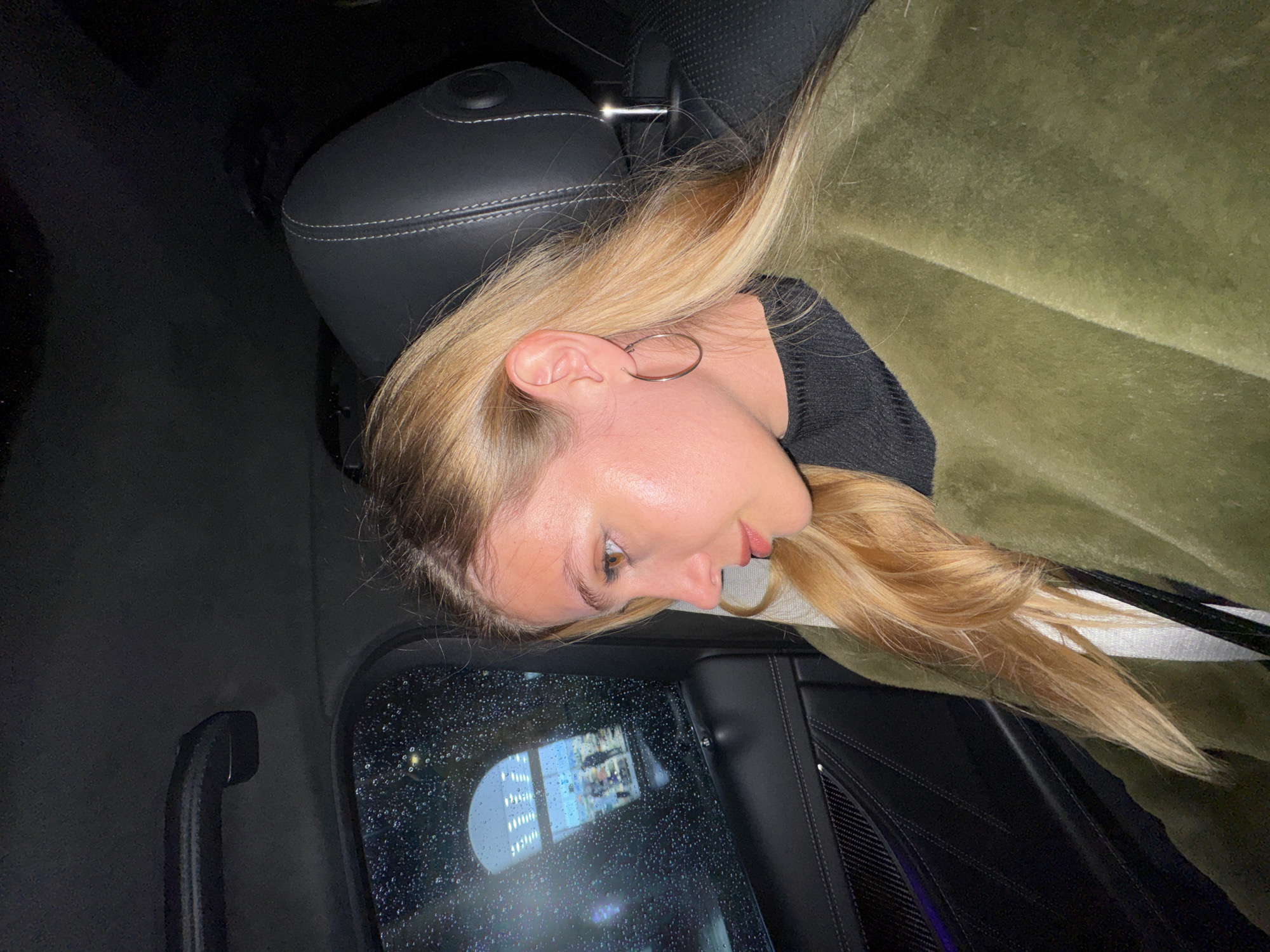 A young woman with long blonde hair is depicted in a close-up, profile shot, seated inside a car. Her head rests against a dark car headrest with visible white stitching. She is wearing a black sweater and a large silver hoop earring is prominent on her visible ear. An olive-green blanket or fabric is draped around her shoulders and behind her.
The setting is a car interior during nighttime or in very low light conditions. The car window beside her is covered in numerous raindrops, indicating rainy weather. Reflections in the wet window show blurred city lights, including a brightly lit building with multiple windows, suggesting an urban environment. A dark grab handle is visible on the car's ceiling or pillar near the window. The woman's expression appears calm and contemplative, as she looks slightly forward and downward. The overall scene conveys a sense of quiet observation from within a vehicle in Tbilisi, Georgia, amidst rain. No text is visible.