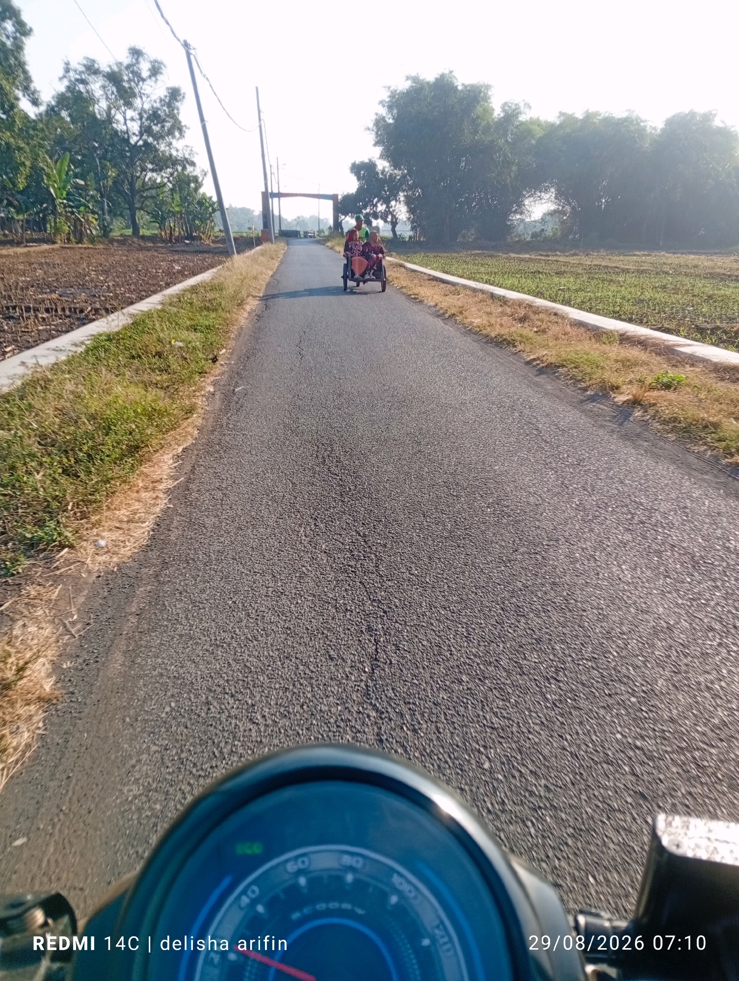 The image captures a sunny day on a paved road in Pukul, Indonesia, with a point-of-view perspective from a moving motorcycle. In the distance, two people are riding in a tricycle-like vehicle, approaching an archway. The road is flanked by fields, one appearing to be recently tilled and the other with young green crops. Trees line the background. The foreground shows a portion of the motorcycle's dashboard, with a speedometer partially visible. The timestamp in the bottom right corner indicates the image was taken on August 29, 2026, at 7:10 AM. Text in the bottom left corner reads "REDMI 14C | delisha arifin".