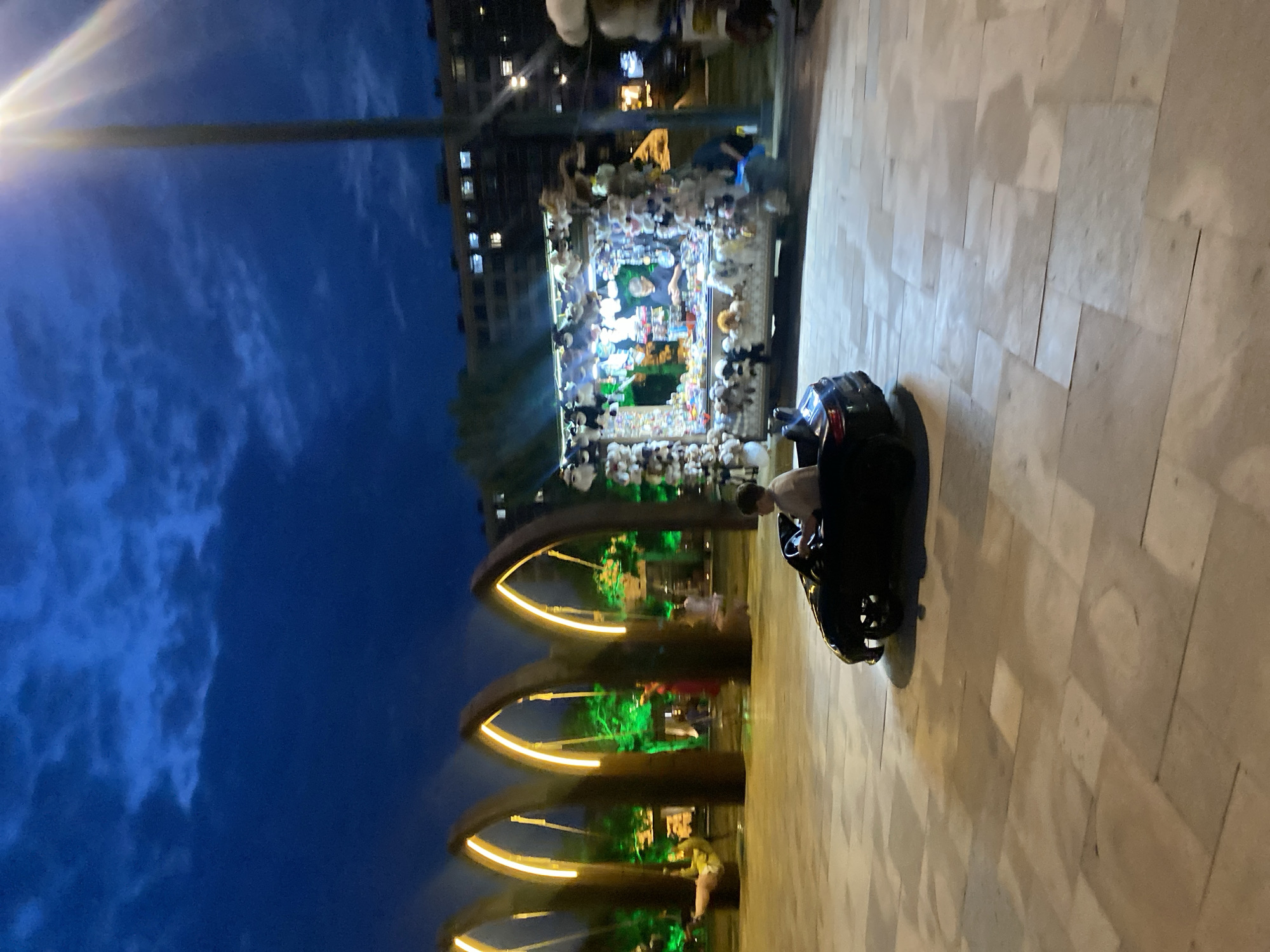 This image depicts an outdoor scene at dusk in Derbent, Russia. The primary subjects are people and various objects displayed in a market stall. The scene is illuminated by artificial lights from the stall, a street lamp, and ambient light from buildings in the background.
In the foreground, a person is seated in what appears to be a toy electric car. The car is black and has visible wheels. The person is wearing a light-colored shirt. The ground is paved with large, rectangular light-colored stones.
The midground features a market stall filled with numerous stuffed animals. The stall is brightly lit, with a variety of plush toys displayed both inside and around it. Behind the stall, there are arched structures with green lighting and illuminated yellow accents. People can be seen moving around this area.
In the background, a multi-story building with illuminated windows is visible. Above the scene, the sky is a deep blue, indicating it is late evening. Wisps of clouds are present in the sky, and a bright street lamp is visible on the left side, casting a beam of light.
The overall impression is that of a lively evening setting, likely a marketplace or public area, where people are enjoying the atmosphere and browsing goods. The presence of the toy car and the abundance of stuffed animals suggest a family-friendly environment. There is no visible text in the image.