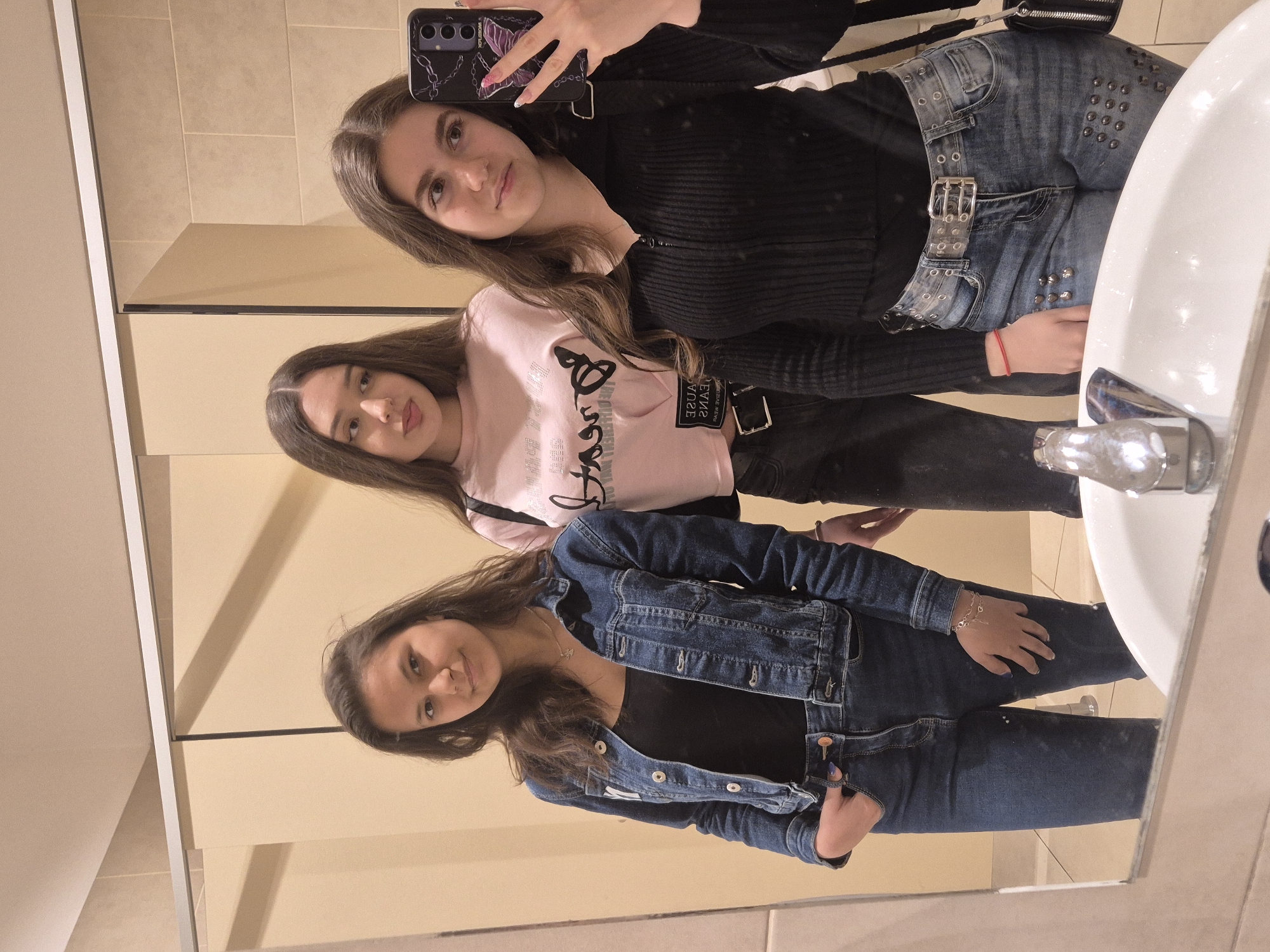 Three young women are captured in a mirror selfie, likely taken in a bathroom in Zagore, Bulgaria. The image is oriented vertically but the subjects appear to be standing upright.
The woman on the left, holding the smartphone, has long brown hair and is wearing a black ribbed long-sleeved top. She is also wearing dark jeans with metal studs or grommets and a wide, transparent belt with multiple silver buckles. Her phone case features a purple butterfly and chain design.
The middle woman has long brown hair and is dressed in a light pink long-sleeved top. Visible text on her shirt, when seen reflected, reads "NO CAUSE DREAMS" in a stylized script. She is wearing dark pants or jeans, and a black strap from a bag or belt displays the mirrored text "CAUSE JEANS".
The woman on the right has long brown hair and is wearing a black top underneath a blue denim jacket with silver buttons. She is dressed in blue jeans and accessorizes with a silver necklace and a delicate silver chain bracelet on her left wrist.
All three women look towards the camera with casual expressions, suggesting a friendly interaction. The background shows light beige tiled walls, typical of a bathroom setting. A white ceramic sink and a chrome faucet are visible on the right side of the reflection. The lighting is artificial, indicating an indoor environment.