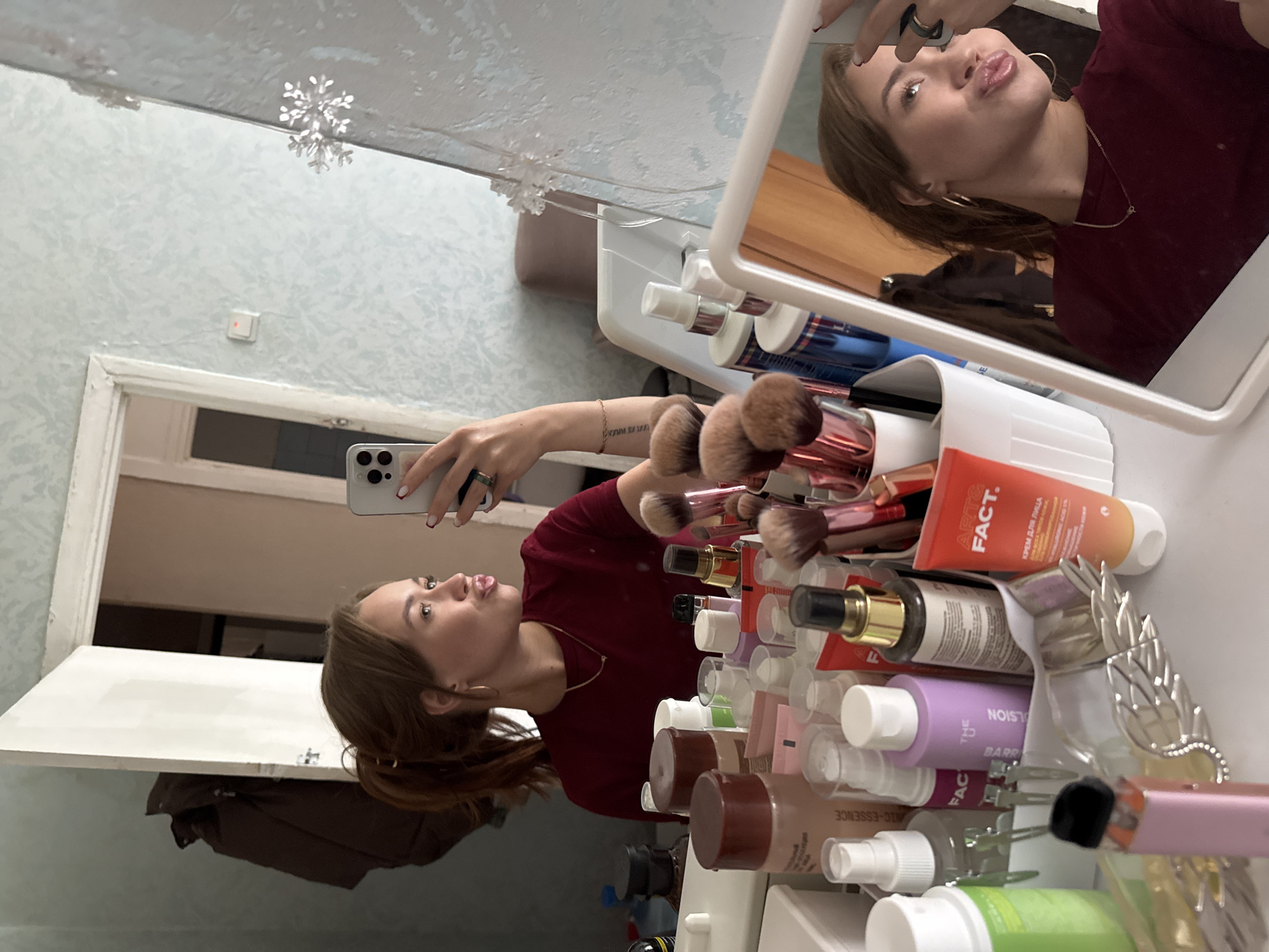 A young woman with brown hair, wearing a maroon t-shirt and gold hoop earrings, is taking a selfie in a mirror. She is making a duck face, sticking her lips out towards the camera. She is wearing a ring on her left hand and a bracelet on her wrist. A tattoo is visible on her arm. She is standing in front of a dresser or vanity, which is filled with various makeup products and brushes. A white mirror is propped up on the vanity, reflecting the woman and the items around her. The background appears to be a room with light blue, textured wallpaper and a white door. A snowflake-shaped decoration is hanging from the mirror or wall. The overall scene suggests the woman is getting ready or has just finished applying makeup. There are several visible brands on the makeup products, including "FACT." and "ESSENCE."