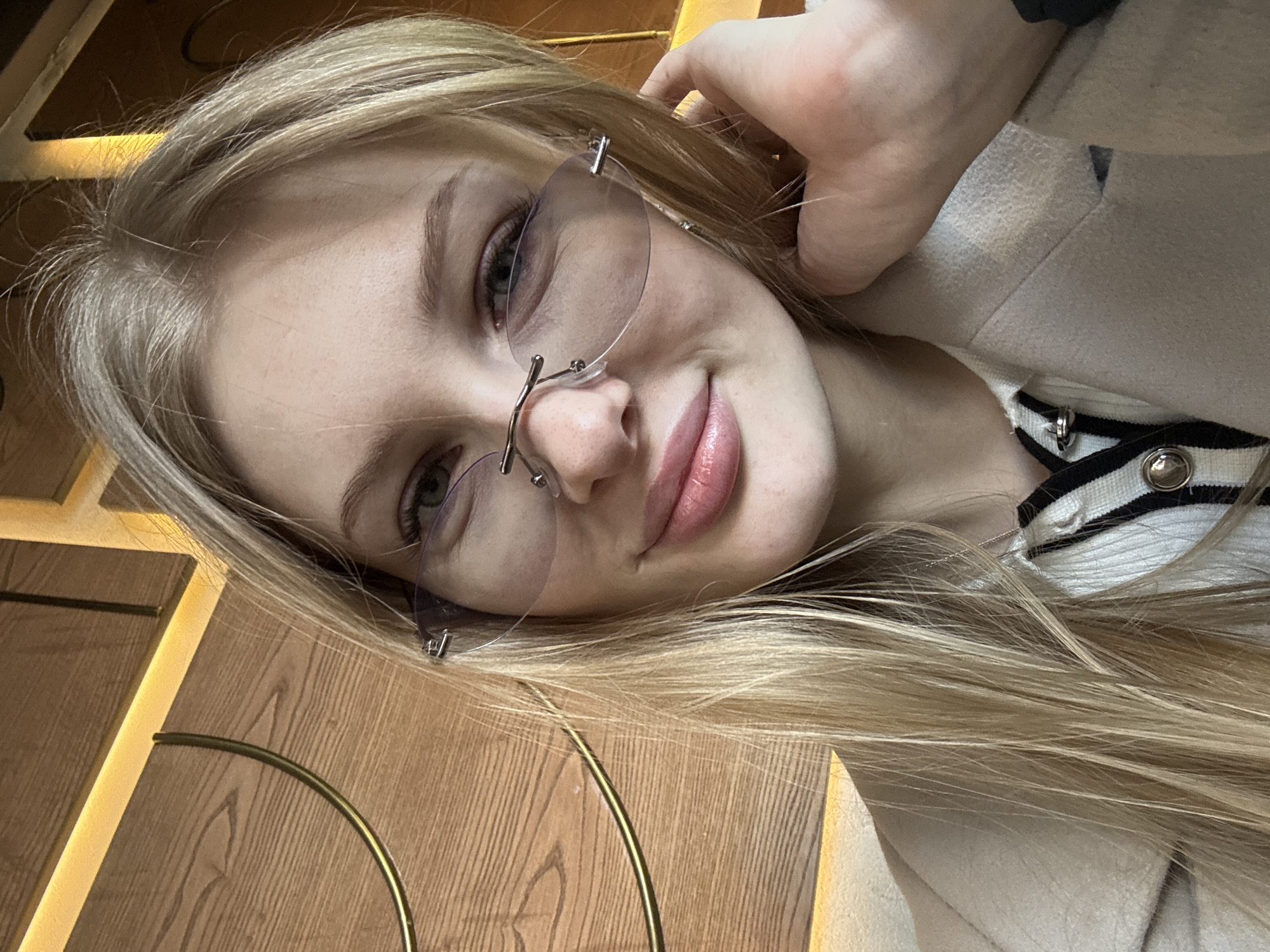 A young woman with long blonde hair and rimless glasses is shown in a close-up, selfie-style photograph. She is wearing a light-colored coat with a striped collar. Her hand is near her head. The background appears to be a wooden surface with decorative gold trim, possibly a table or wall panel. Given the prompt mentioning the city of Perm, Russia, it's reasonable to infer that the photograph was taken somewhere in Perm, although specific details to confirm the location are not visible in the image.