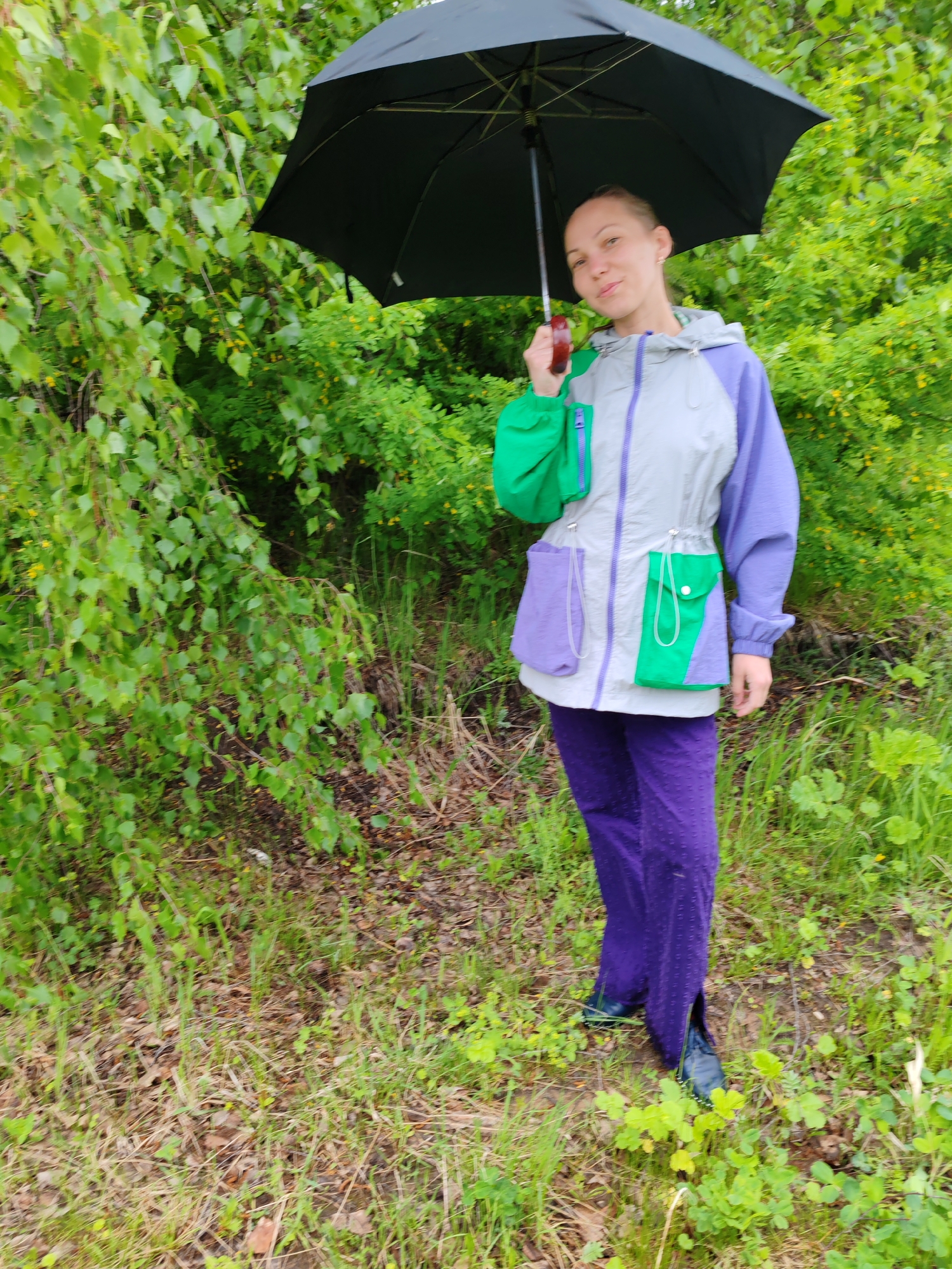 In the city of Чухлэм, Russia, a woman stands in a grassy area with trees and foliage visible in the background. She is holding a black umbrella over her head. The woman is wearing a gray jacket with green and purple color-blocked sections, purple pants with textured dots, and dark shoes. The ground around her is covered in grass and scattered leaves.