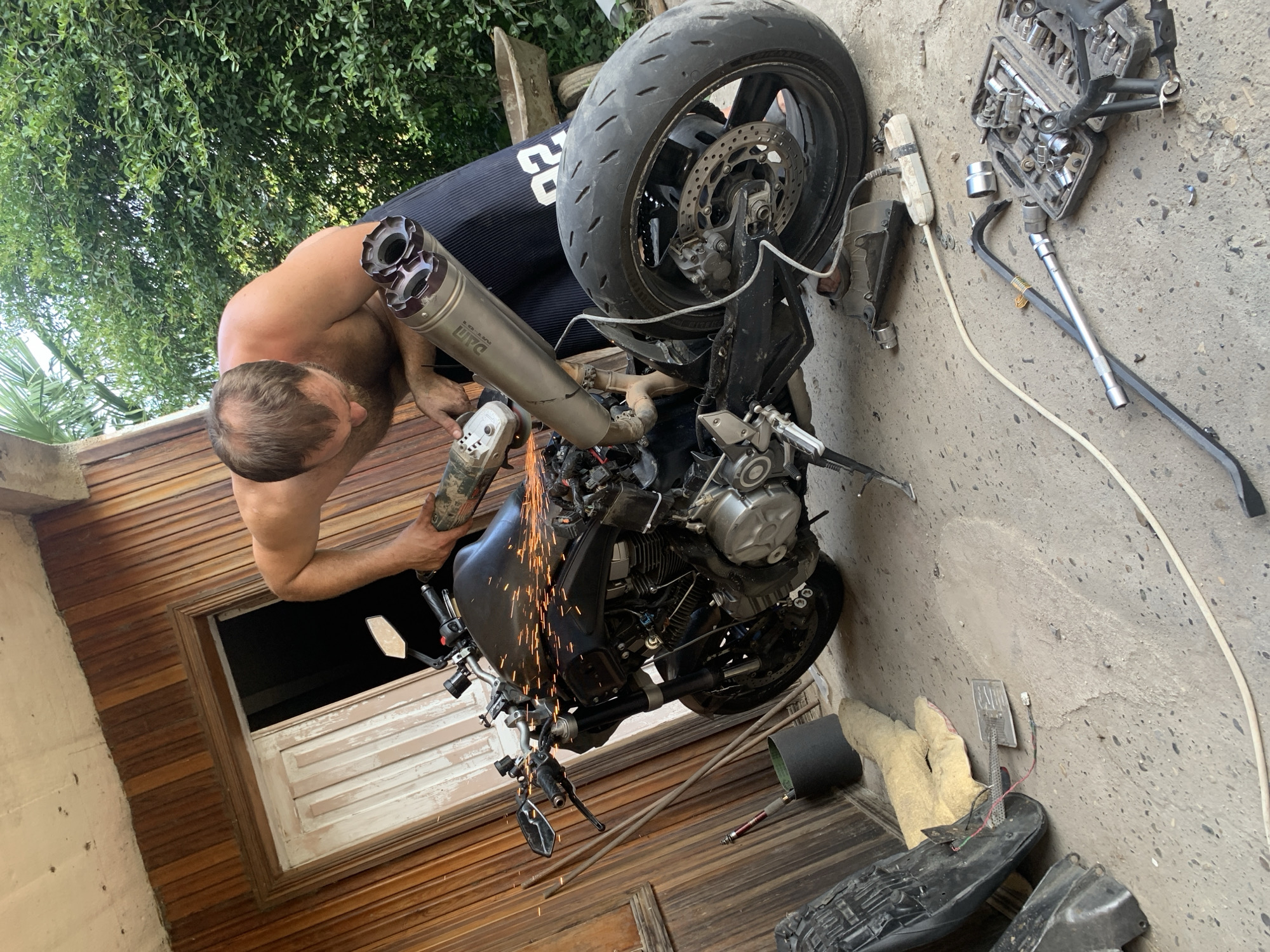 A shirtless man with brown hair is working on a black motorcycle, using an angle grinder that is emitting sparks. He is wearing dark shorts with a white number '02' on the side. The motorcycle is partially disassembled, with the exhaust system removed and visible. Tools are scattered on the concrete ground around the motorcycle, including a toolbox filled with various wrenches and sockets. The scene appears to be outdoors, possibly in a yard or driveway, with wooden siding and greenery in the background. A wheelbarrow is visible in the upper right corner. The man is focused on the task, with his body positioned over the motorcycle's engine.