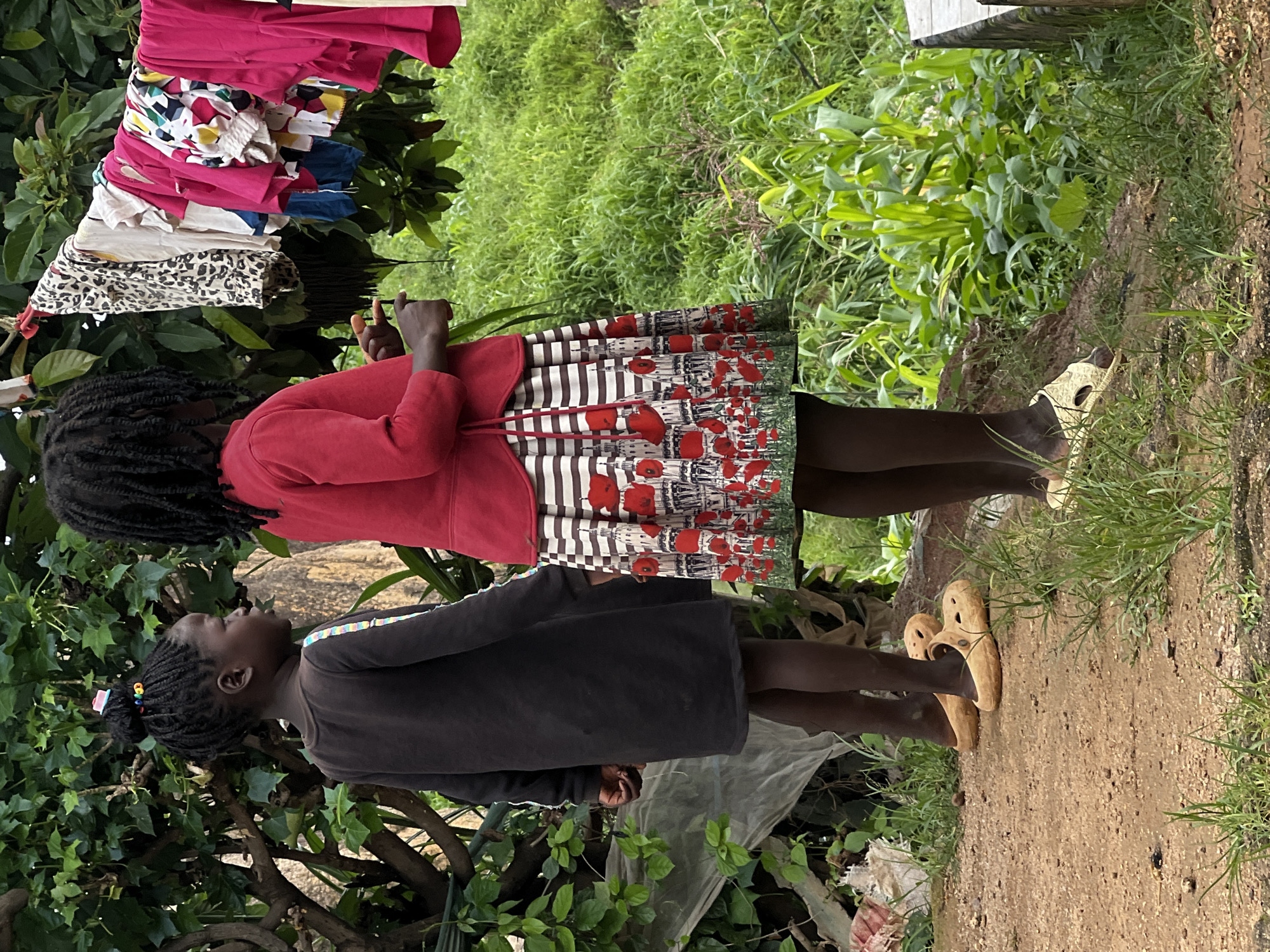 In Jos, Nigeria, two young girls stand outdoors near lush green vegetation. The girl on the left wears a dark long-sleeved dress and tan clogs, her hair styled in braids with colorful beads. The girl on the right is dressed in a red jacket and a skirt with a white and brown striped pattern adorned with red poppy and building motifs. Her dark braided hair is visible. Behind them, laundry hangs on a clothesline under a leafy canopy. The ground is dirt with patches of grass, and a white plastic sheet is draped over some unseen objects to the left. The scene is set against a backdrop of dense green foliage and tall plants, possibly corn stalks.