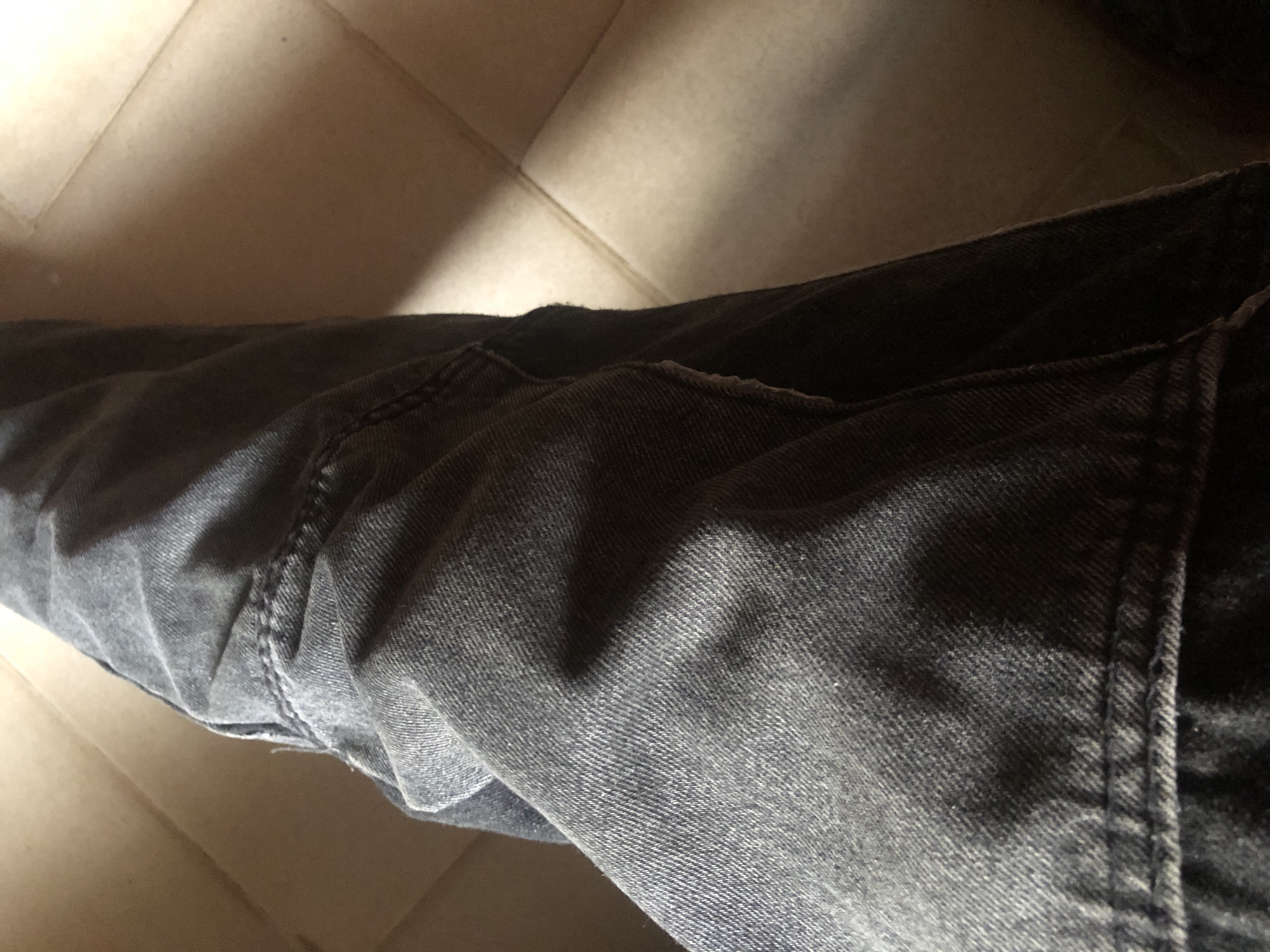 The image depicts a close-up view of what appears to be a person's leg clad in gray denim jeans. The fabric is textured with visible stitching and creases. The background is a tiled floor, with tiles in shades of beige. The location is unknown.
