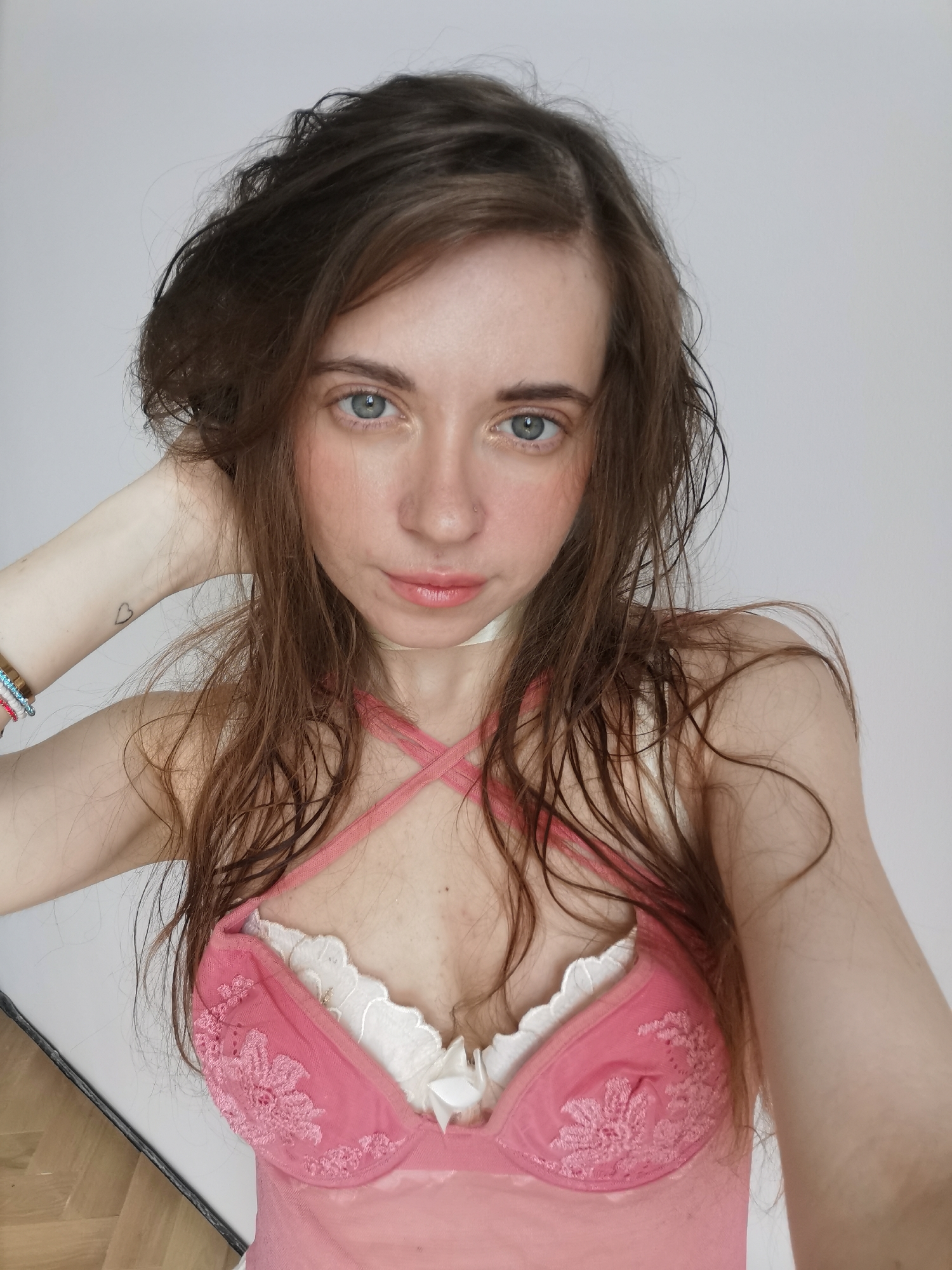 A young woman is posing indoors. She has fair skin, brown hair, and blue eyes. She is wearing a pink and white lingerie top. Her left hand is raised behind her head. A small heart tattoo is visible on her left wrist, along with beaded bracelets. The background is a plain white wall. There are no indications in the image that suggest Moscow, Russia as a location.