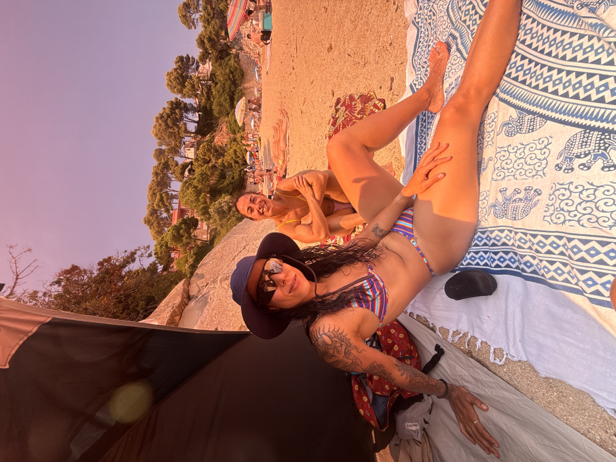 Two women are sunbathing on a sandy beach in Calonge i Sant Antoni, Spain. The woman in the foreground, wearing a striped bikini and a blue hat, is lying on a blanket with her legs extended. She has tattoos on her arms and is wearing sunglasses. The woman in the background, wearing a yellow bikini, is sitting with her arms crossed. They are near a black tent. The beach is populated with other people and beach umbrellas in the background. The landscape behind the beach features rocky hills covered with pine trees and some buildings. The sky is clear and a light purple hue.