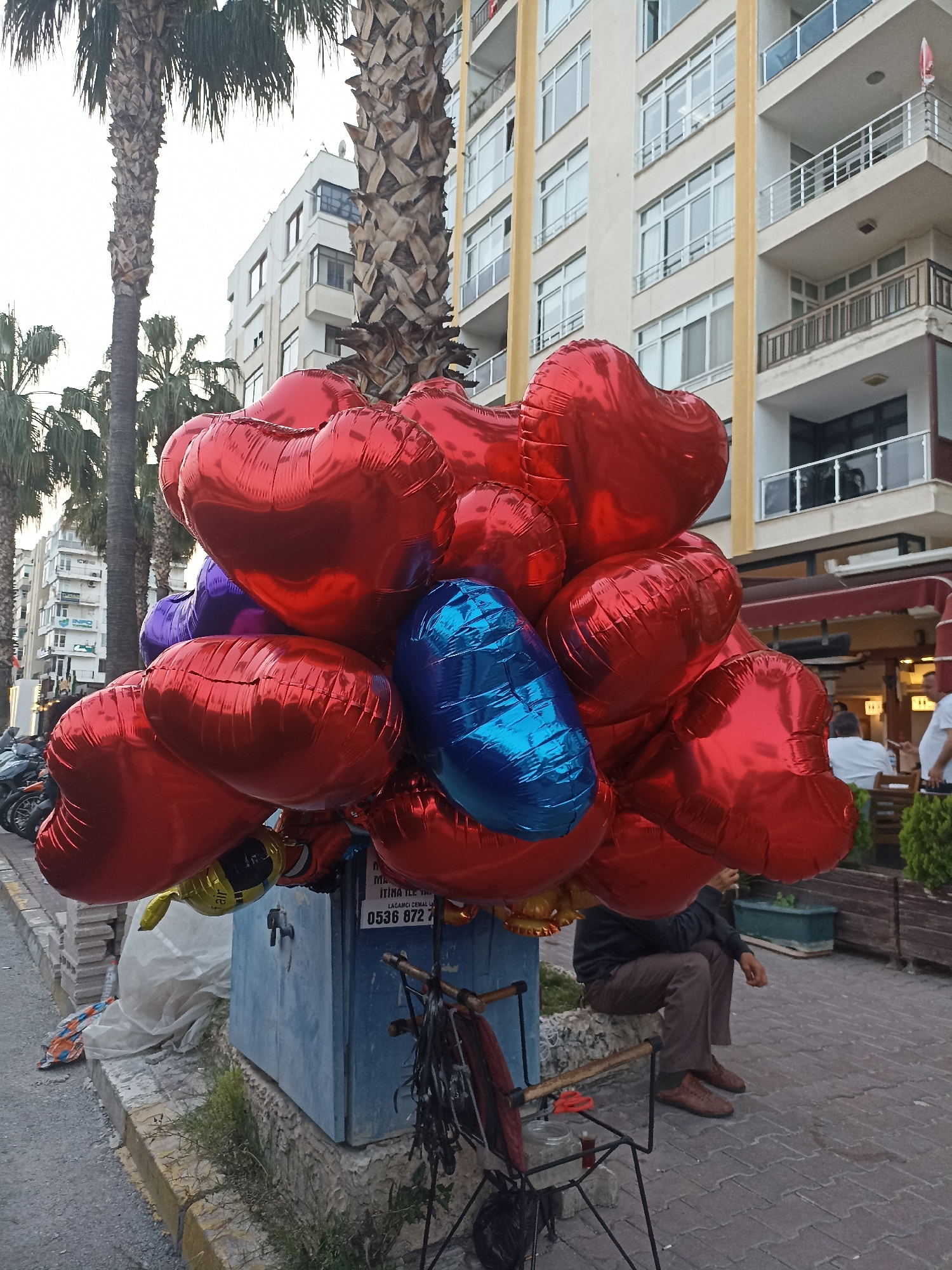 The image shows a collection of heart-shaped balloons, mostly red with some blue and purple ones mixed in. The balloons are bunched together and attached to a blue metal box. A bicycle frame is leaning against the box. A man is sitting on a low concrete wall near the balloons, wearing brown pants, a dark jacket, and brown shoes. In the background are buildings and palm trees. People are sitting at tables in what appears to be an outdoor cafe. The scene is located in Yenişehir, Turkey, evidenced by the phone number on the metal box: 0536 872.