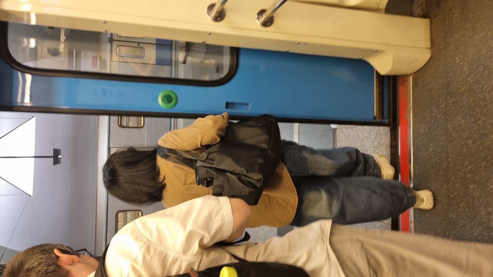 A woman with dark hair, wearing a tan button-down shirt and blue jeans, walks towards an open subway door on the platform in Moscow, Russia. She carries a black tote bag over her left shoulder. To her left, a person wearing a light-colored short-sleeved shirt with a backpack is partially visible. The subway car has a blue door with a green button. The floor of the platform is dark grey with a red strip near the door opening.