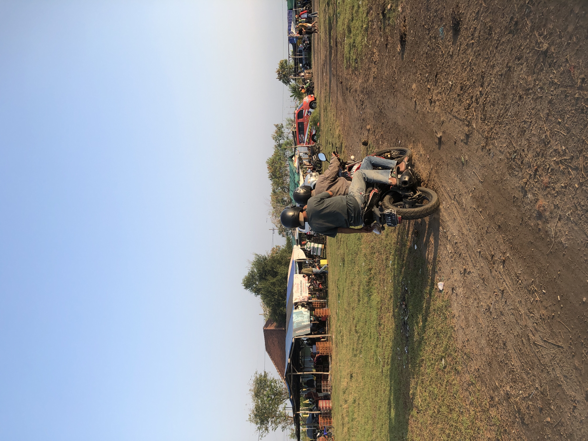 In Gempolsari, Indonesia, two individuals are seen riding a motorcycle down a dirt path. The person in the back is wearing a dark t-shirt and jeans, while the driver is wearing a light brown jacket and jeans. Both are wearing helmets. The motorcycle has a license plate visible. The surrounding environment is an open, grassy area with scattered dirt patches. In the background, a marketplace is set up with several stalls, some covered by tarps, and various goods displayed in crates. Several vehicles, including cars and other motorcycles, are parked around the marketplace. Trees and vegetation are visible in the distance, and the sky is clear and light blue. The scene appears to be a daytime outdoor market in a semi-rural setting.