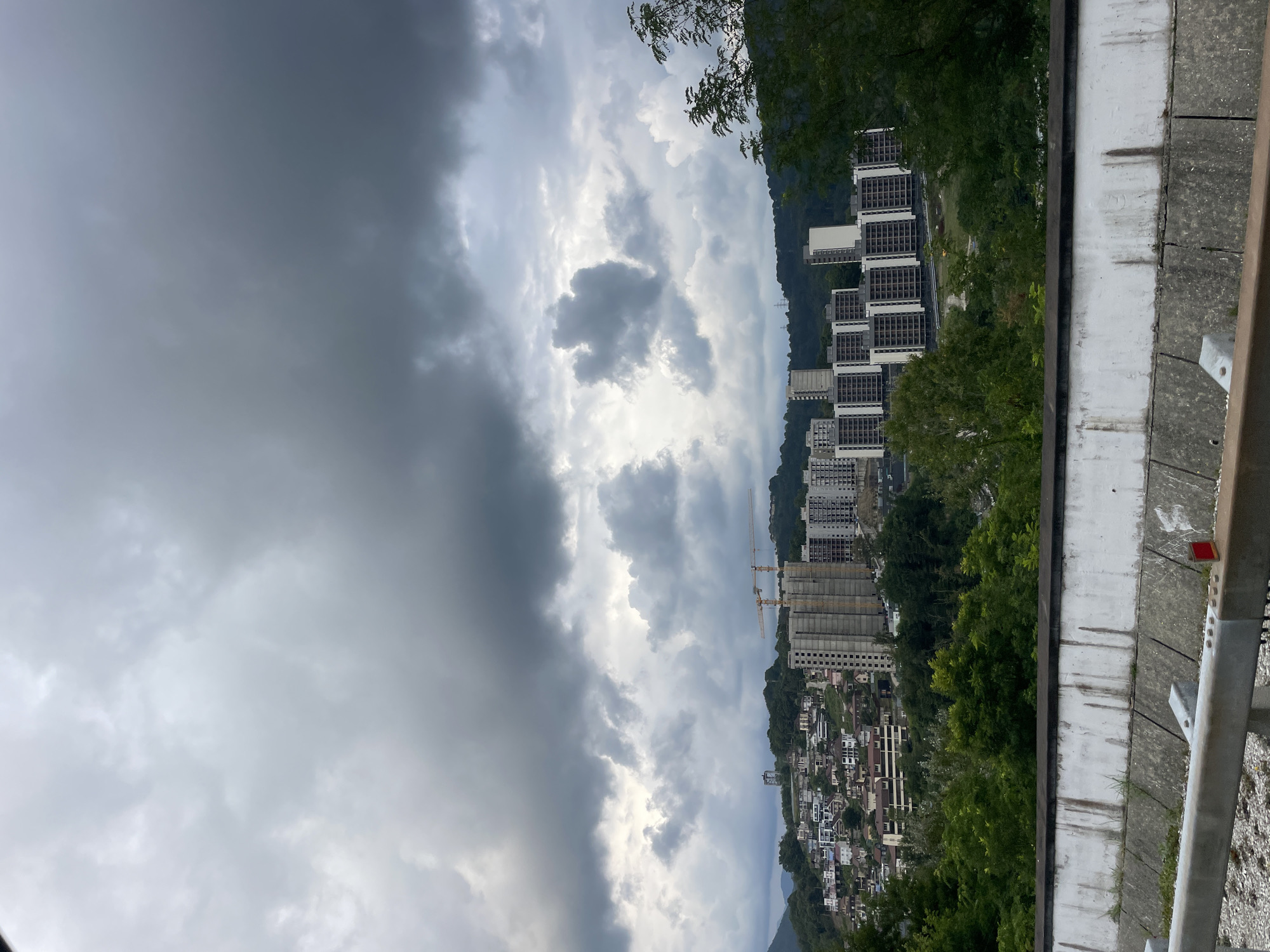 The image shows a view of a hillside urban area in Sochi, Russia, under a heavy, overcast sky. The foreground features a concrete barrier and a metal railing, likely part of a roadside overlook or highway. Beyond this, a densely populated hillside is visible, with many multi-story residential buildings and houses climbing the slope. Construction is evident, with at least two tall, unfinished apartment buildings and construction cranes situated in the middle ground. Lush green trees and vegetation cover the lower slopes and dot the landscape between the buildings. The background consists of rolling, forested hills and mountains. The overall atmosphere is somber due to the dark, dramatic cloud cover.