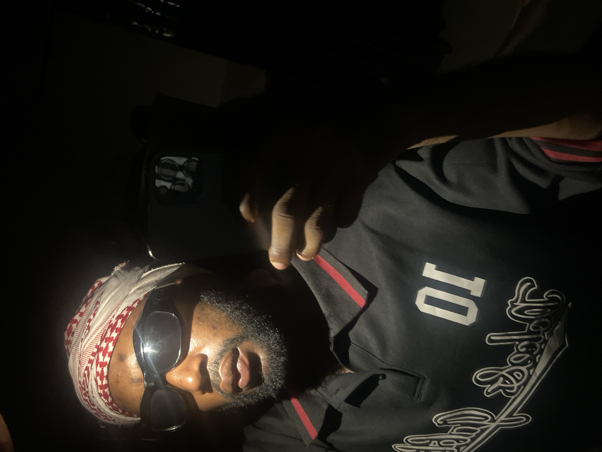 A person is seen wearing a headscarf and sunglasses. The person is also wearing a black shirt with the number "10" and a logo on it. A hand holding a phone is visible. The background is dark. It is not possible to identify the location of Lokoja, Nigeria from the provided image.