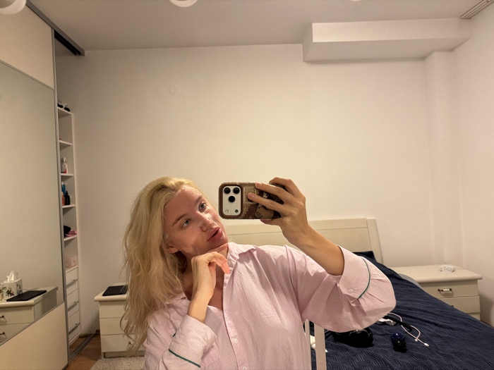 A female individual with long blonde hair is depicted taking a mirror selfie in a bedroom located in Budva, Montenegro. She is wearing a light pink, striped long-sleeved shirt with green piping on the cuffs. Her face appears smooth, possibly from a facial mask or moisturizer, and she holds a smartphone with a camouflage-patterned case in her right hand, positioned to capture her reflection. Her left hand rests beneath her chin.
The bedroom features a bed with a white headboard and a dark blue comforter. Matching white nightstands are positioned on either side of the bed. The left nightstand, partially obscured by the individual's reflection, displays a tissue box and a dark, flat electronic device. The right nightstand holds a white object and a charging cable. On the dark blue comforter, near the foot of the bed, a black pouch, white wired earphones, and a small dark blue object are present.
To the left of the frame, a large light-colored wardrobe with a mirrored sliding door reflects an internal shelving unit stocked with various bottles and containers. The walls of the room are white, and a rectangular white protrusion is visible on the ceiling in the upper right area. The floor in the foreground is covered by a light-colored carpet.