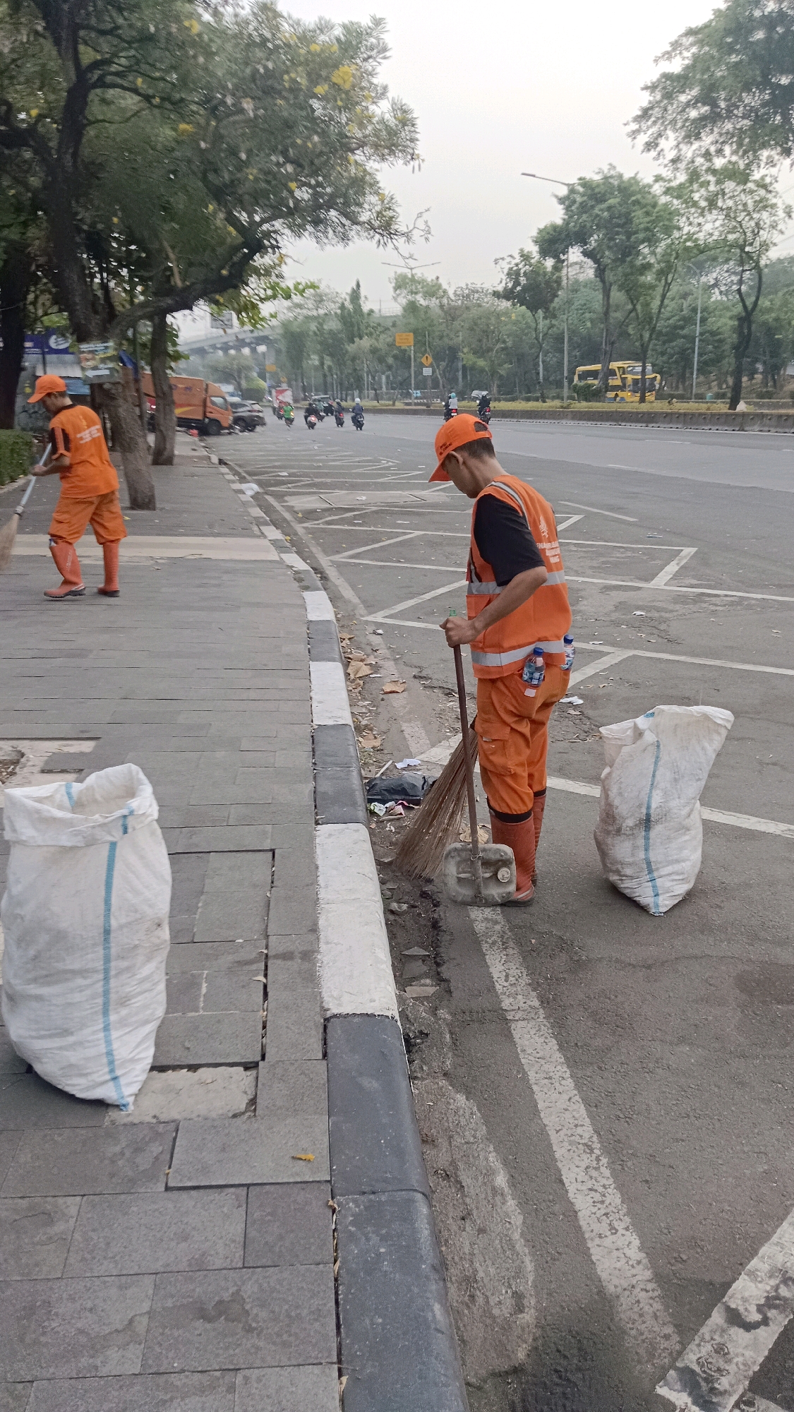 Two sanitation workers are visible on a sidewalk next to a road. The worker on the left, in the foreground, is wearing an orange uniform, cap, and boots, and is sweeping the sidewalk with a broom. A large white sack is on the sidewalk to the left of this worker. The worker on the right, in the middle ground, is also in an orange uniform, cap, and boots, and is sweeping the gutter with a broom. Another white sack is on the asphalt road to the right of this worker. Several vehicles, including motorcycles, cars, and trucks, are on the road in the background. Trees line the side of the road. The scene appears to be in a public area, likely a street in East Jakarta, Indonesia, given the presence of sanitation workers in uniforms and the typical urban environment. The sky is overcast, suggesting it might be early morning or late afternoon.