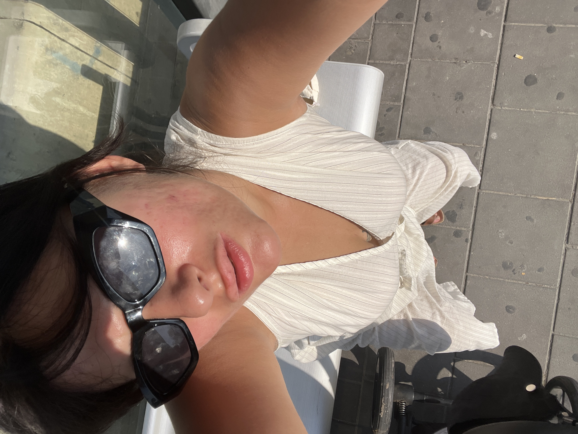 A woman is taking a high-angle selfie outdoors, under bright sunlight. She is wearing black, angular sunglasses and a white, sleeveless, ribbed dress with a V-neckline. Her skin appears flushed and oily, with some redness and blemishes visible on her cheeks and forehead. She has dark hair, some of which is falling across her face. Her lips are slightly parted, revealing a glossy sheen.
In the background, to her right, is a modern, silver bench with a ribbed texture. To her left and slightly in front of her, is a portion of a black baby stroller. The ground beneath her is paved with large, dark grey square tiles, some of which have circular stains. There is a discarded cigarette butt on the pavement. The overall setting appears to be an urban outdoor public space, possibly near a bus stop or a park, in Kiryat Motzkin, Israel.