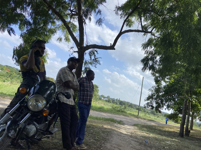 Three men stand beneath a tree next to a motorcycle. One man sits on the bike, talking on a cell phone. Another wears a plaid shirt and a baseball cap, with arms crossed. The third man wears a red and blue plaid shirt and blue jeans. A dirt road extends into the distance where another person walks. The sky is partly cloudy.