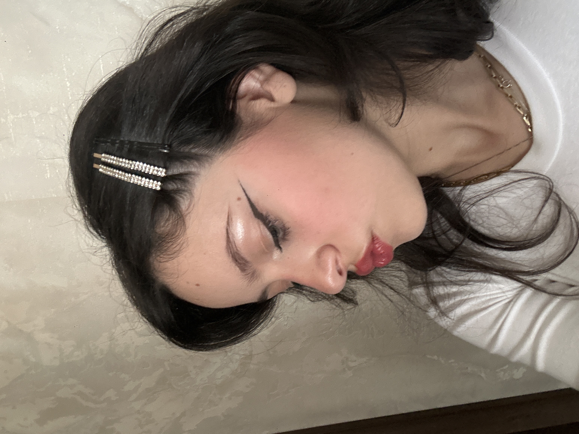 A person lies on a beige surface, possibly a bed, with dark hair and a hair clip. They have a gold chain visible on their neck, visible eyeliner on their closed eyes, and pink blush on their cheeks. The person is wearing a white shirt. The image does not provide any information that allows for the determination of a location in Yerevan, Armenia or otherwise.