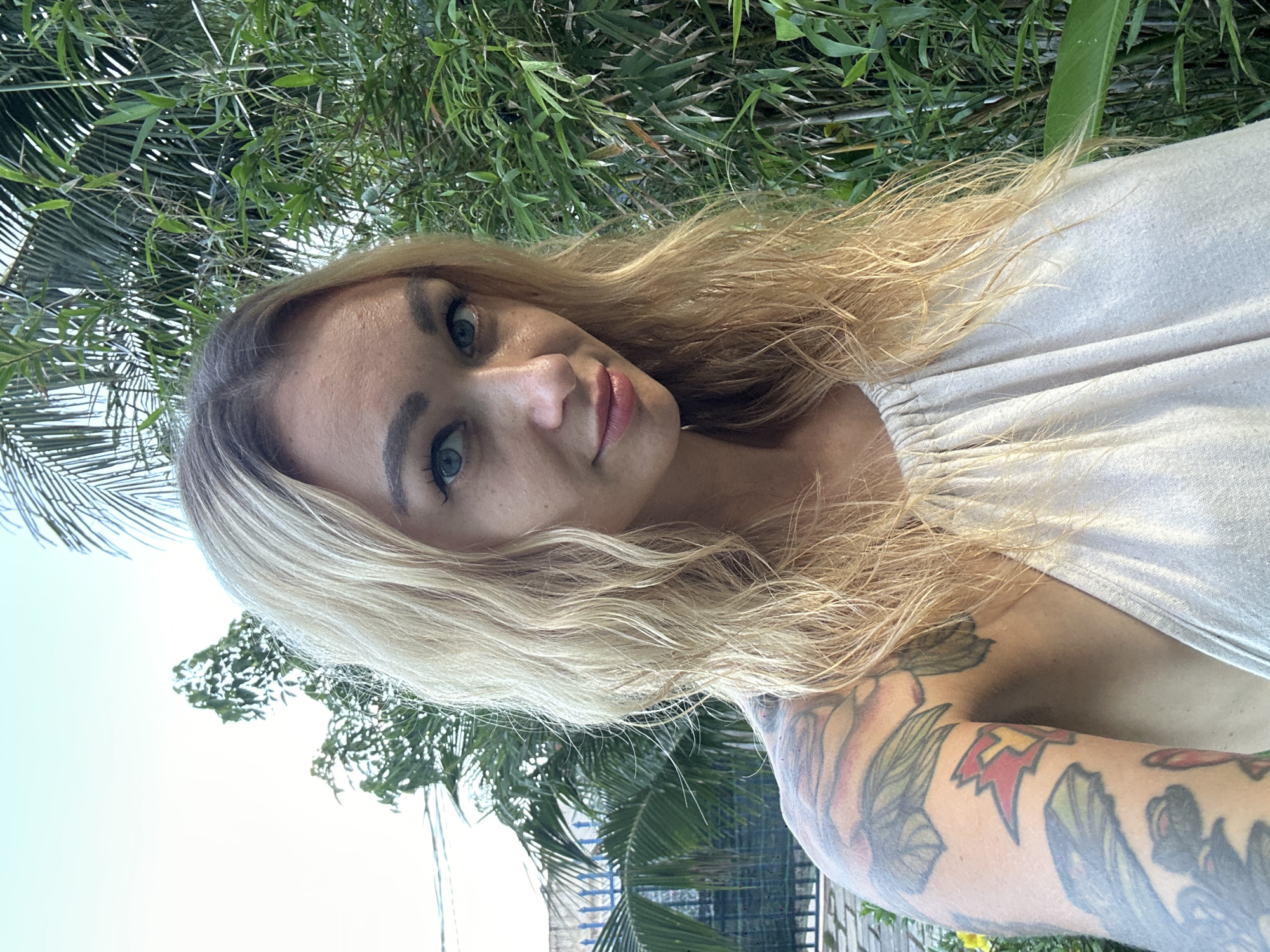 An outdoor portrait captures a woman with long, wavy blonde hair and striking blue eyes, looking directly at the camera with a subtle, calm expression. Her eyebrows are dark and well-defined, and she wears light lipstick. She is dressed in a simple, light-colored, possibly beige, sleeveless top. Her right arm is heavily adorned with colorful tattoos, featuring various designs that include floral motifs and a distinct red lightning bolt symbol with a yellow accent.
The background is dominated by lush green foliage, with visible palm fronds and other leafy tropical plants, indicating a warm, natural environment. Glimpses of a clear, bright blue sky are visible through the leaves, suggesting it is daytime with sunny weather. The image appears to be a close-up selfie or portrait taken in an outdoor setting, consistent with the tropical climate of Mũi Né, Vietnam. There is no visible text in the image.