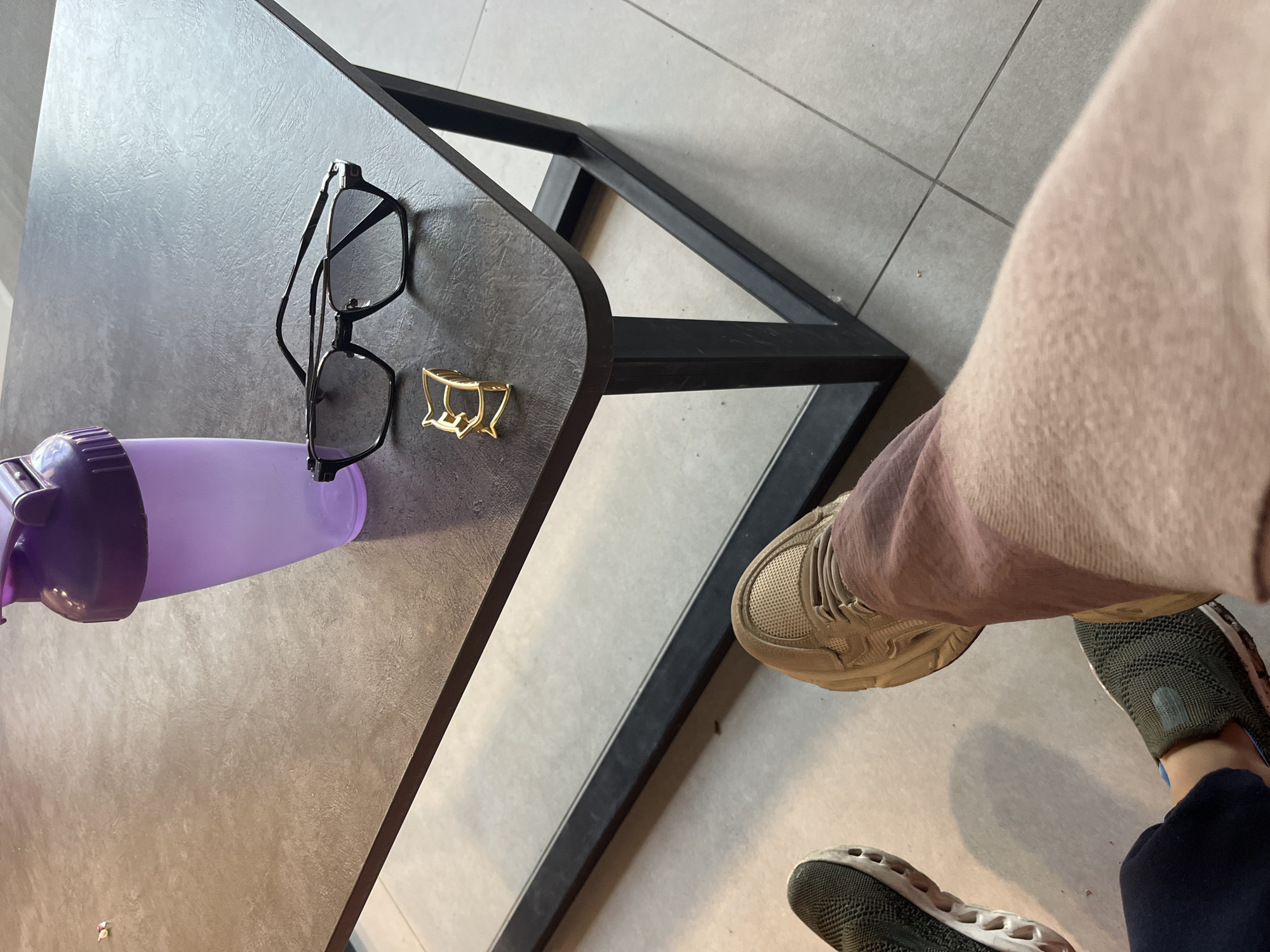 The scene appears to be indoors, on a tiled floor in Kaspiysk, Russia. A dark-colored, square table with rounded corners is the central focus. On the table are a purple water bottle, a pair of black-framed eyeglasses, and a gold-colored hair clip. A person wearing light brown pants and sneakers is partially visible to the right of the table. The person's feet are resting on the floor. The floor is tiled with light grey square tiles.