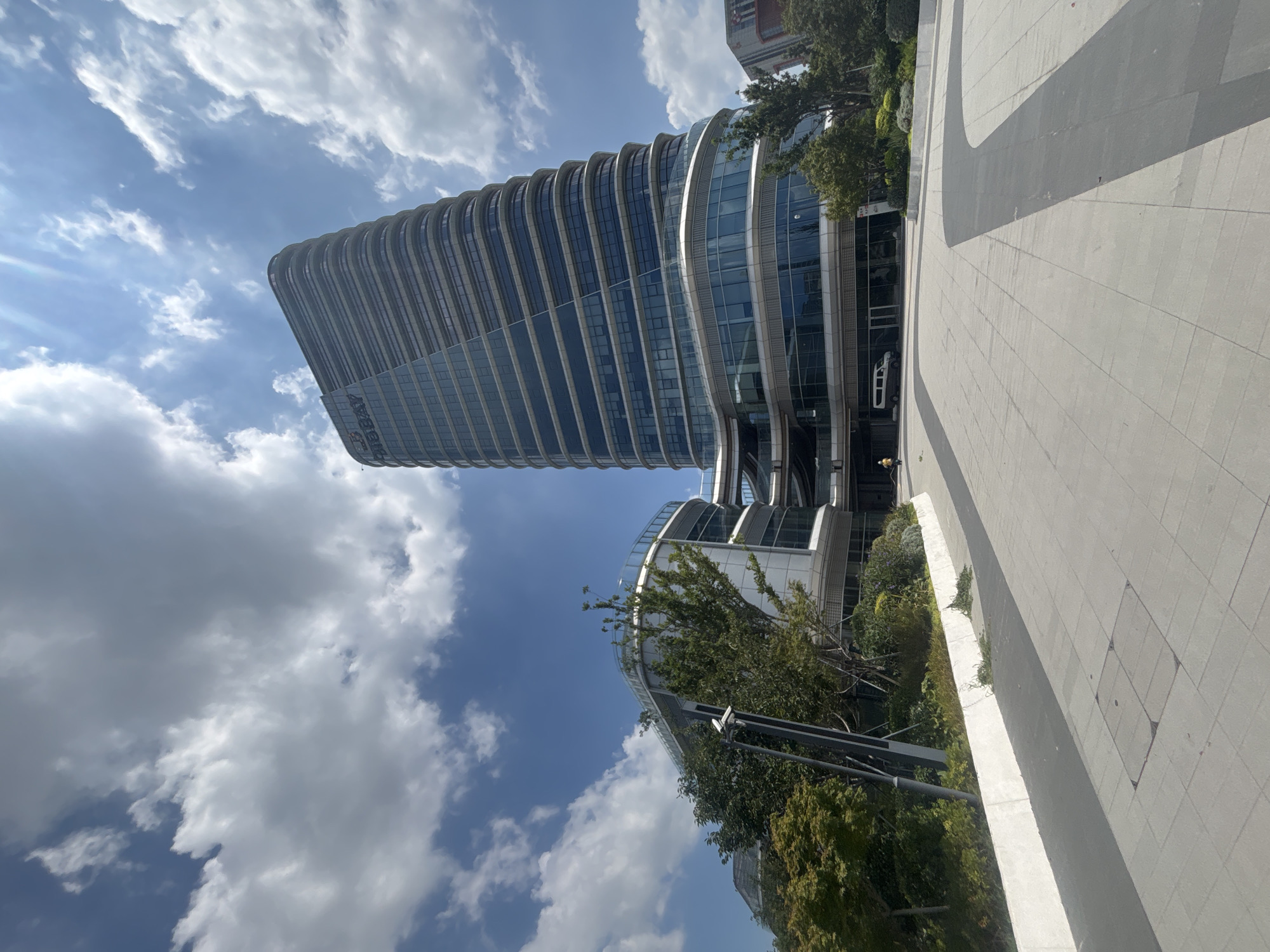 A wide-angle, low-angle exterior shot captures a modern, curved skyscraper and an adjacent, lower, curved building on a sunny day with scattered clouds. The skyscraper, identifiable by the "BIGBAY" logo on its upper portion, features a glass facade with horizontal banding that creates a wavy effect. The lower building, also with a glass facade, is partially obscured by lush green trees and shrubs. A paved walkway with distinct grey and light grey sections leads towards the buildings, with a white planter box running alongside the walkway. In the background, a white van is parked near the entrance of the lower building, and a person in yellow clothing is walking in the distance. The scene is set in the Suzhou Industrial Park, China.