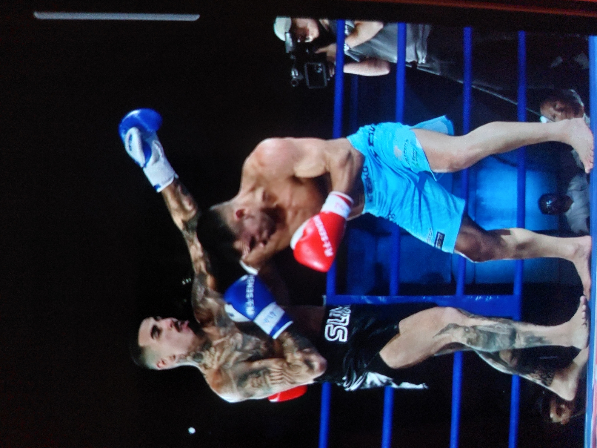 This image captures an intense moment during a combat sports event, likely boxing or kickboxing, taking place in a ring in Tsarevo, Bulgaria. The scene is illuminated by artificial lighting, suggesting an evening or night event.
Two male fighters are actively engaged in the center of the ring. One fighter, with extensive tattoos covering his arms, chest, and neck, and sporting a short, dark haircut, is wearing black shorts with white text that appears to read "SLA". He is also wearing blue and white boxing gloves, with partial text "RINGSID-" visible on one glove. This fighter is depicted in the act of throwing or having just thrown a punch, with his right arm extended towards his opponent. His face is visible, showing a look of exertion with his mouth open.
His opponent, who has fewer visible tattoos, is wearing light blue shorts and red and white boxing gloves. Partial text "RINGSIDE" is visible on one of his red gloves. He is leaning forward, appearing to either absorb a strike, defend, or move into an attack. His left arm is raised, and his face conveys focus or strain.
The fighters are contained within the combat area defined by blue vertical ropes and posts of the ring. In the background, beyond the ropes, several individuals are visible, including what appears to be a photographer holding a camera, suggesting professional coverage of the event. The dark background emphasizes the action within the brightly lit ring, highlighting the physical intensity of the competition.