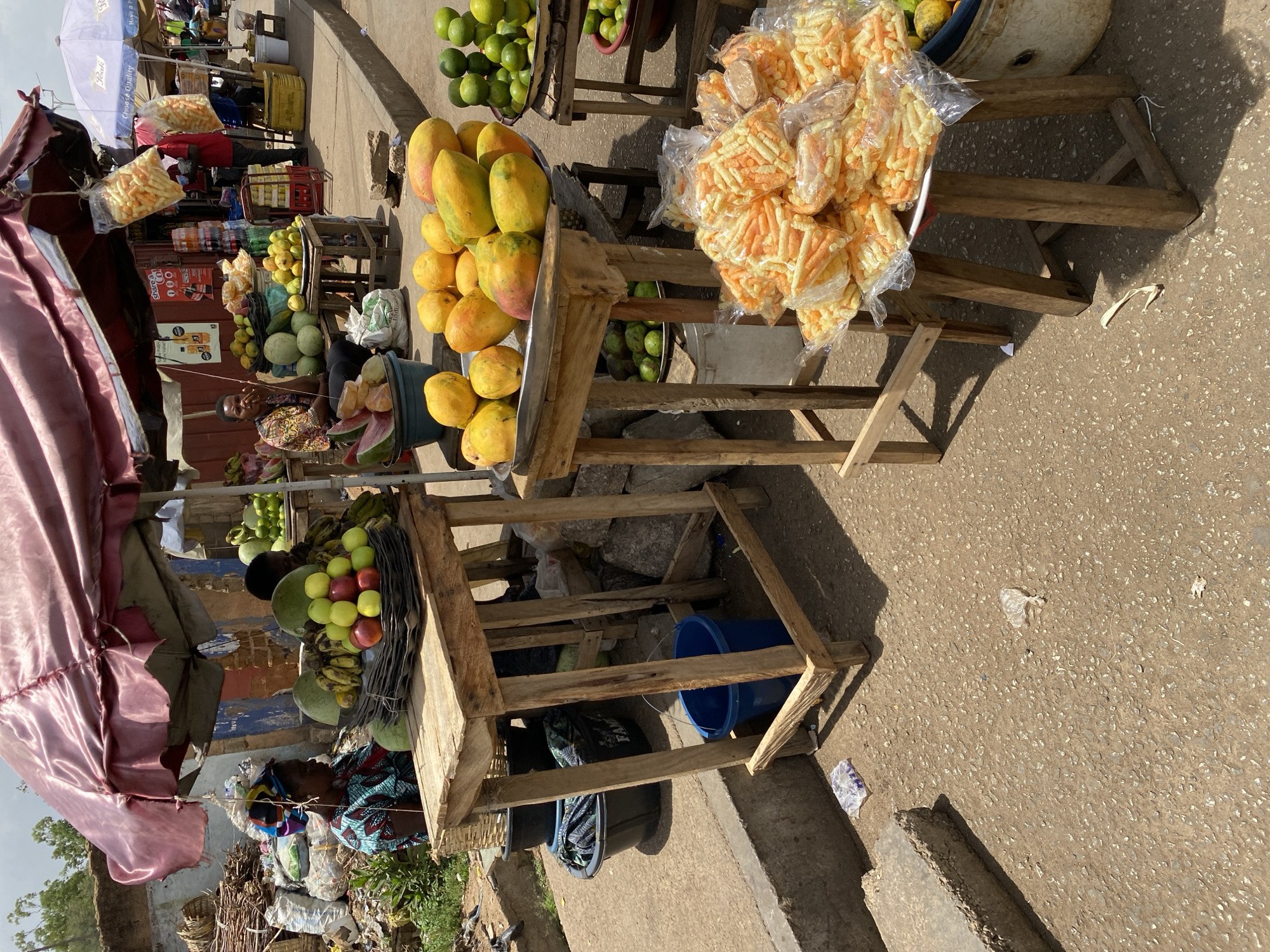 This image captures a vibrant outdoor market scene in Bukuru, Nigeria, bustling under bright midday sunshine. Numerous makeshift stalls, constructed from rough wooden planks, line a coarse, light brown pathway. These stalls are laden with fresh produce and packaged goods.
In the foreground, large quantities of ripe mangoes (yellow and orange) are displayed in metal trays, alongside piles of green limes or oranges. Another prominent stall features numerous clear plastic bags filled with orange and light yellow puffed snacks. Further back, a stall offers bottled beverages, including Coca-Cola, Fanta, and local brands like "Sharpa Cola" and "Big Cola," visible on crates and signage. Other fresh produce includes watermelons, green apples, red apples, and bananas.
A woman, likely a vendor, is visible sitting behind one of the stalls in the lower left, wearing a colorful patterned dress, with cut watermelons in a blue bucket beside her. Another person in a red shirt is partially visible deeper within the market. Canopies and umbrellas provide shade, including a maroon fabric canopy in the foreground and a light blue umbrella with "Quality" partially visible on it. Strong shadows are cast on the ground, indicating the time of day. The market appears lively and informal, with various goods displayed for sale.