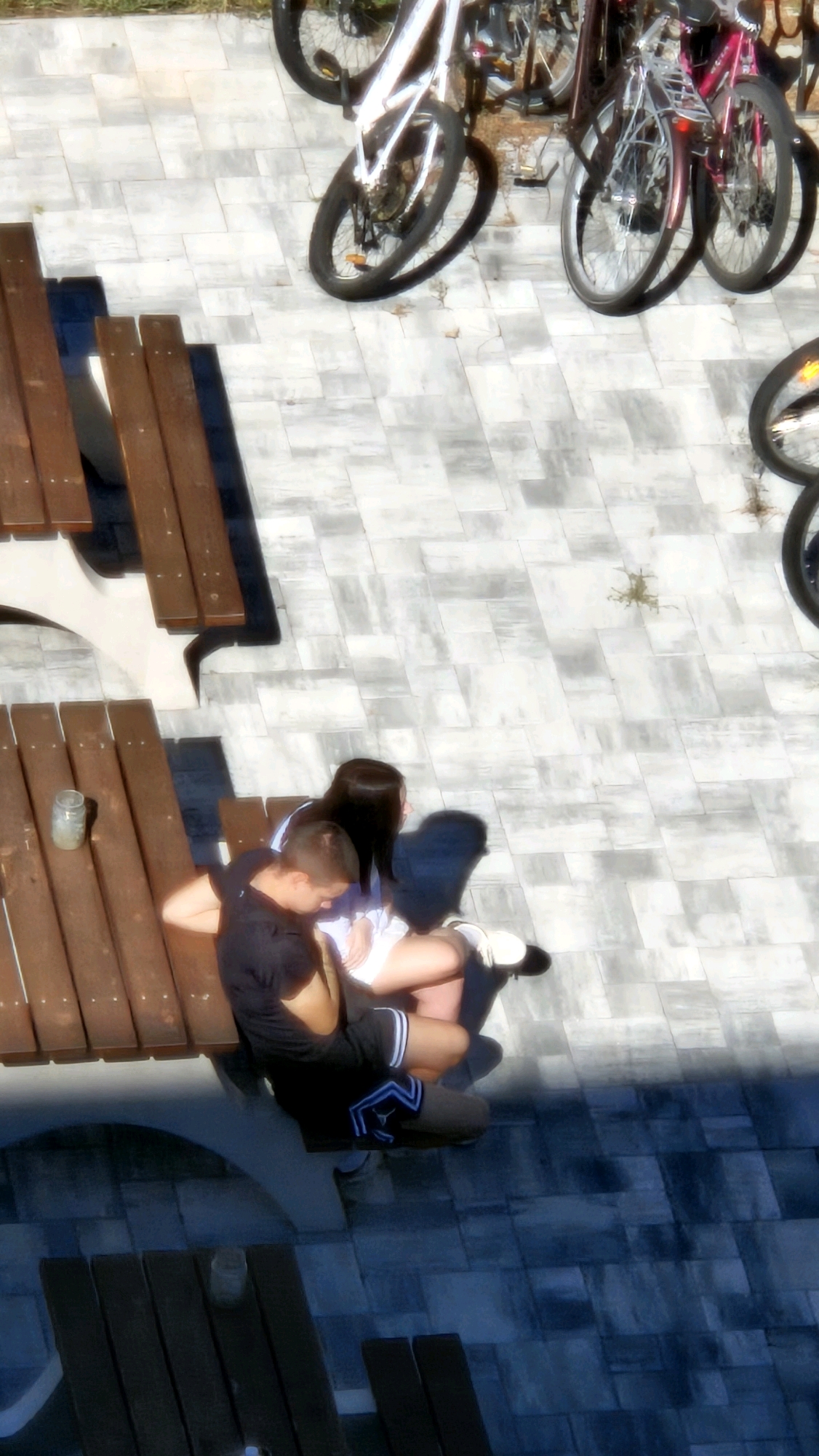 The image shows a top-down view of an outdoor area with a tiled floor. Several bicycles are parked on the right side of the image. In the center-left, a wooden picnic table is visible. On the picnic table, a young man in a black t-shirt and shorts is sitting with his arm around a young woman who is wearing a white top and shorts. They are positioned as if embracing or cuddling on the bench of the table. The lighting suggests it is daytime, with shadows cast on the tiled ground. The overall scene appears to be a public space, possibly a park or a courtyard in the city of Prešov, Slovakia.