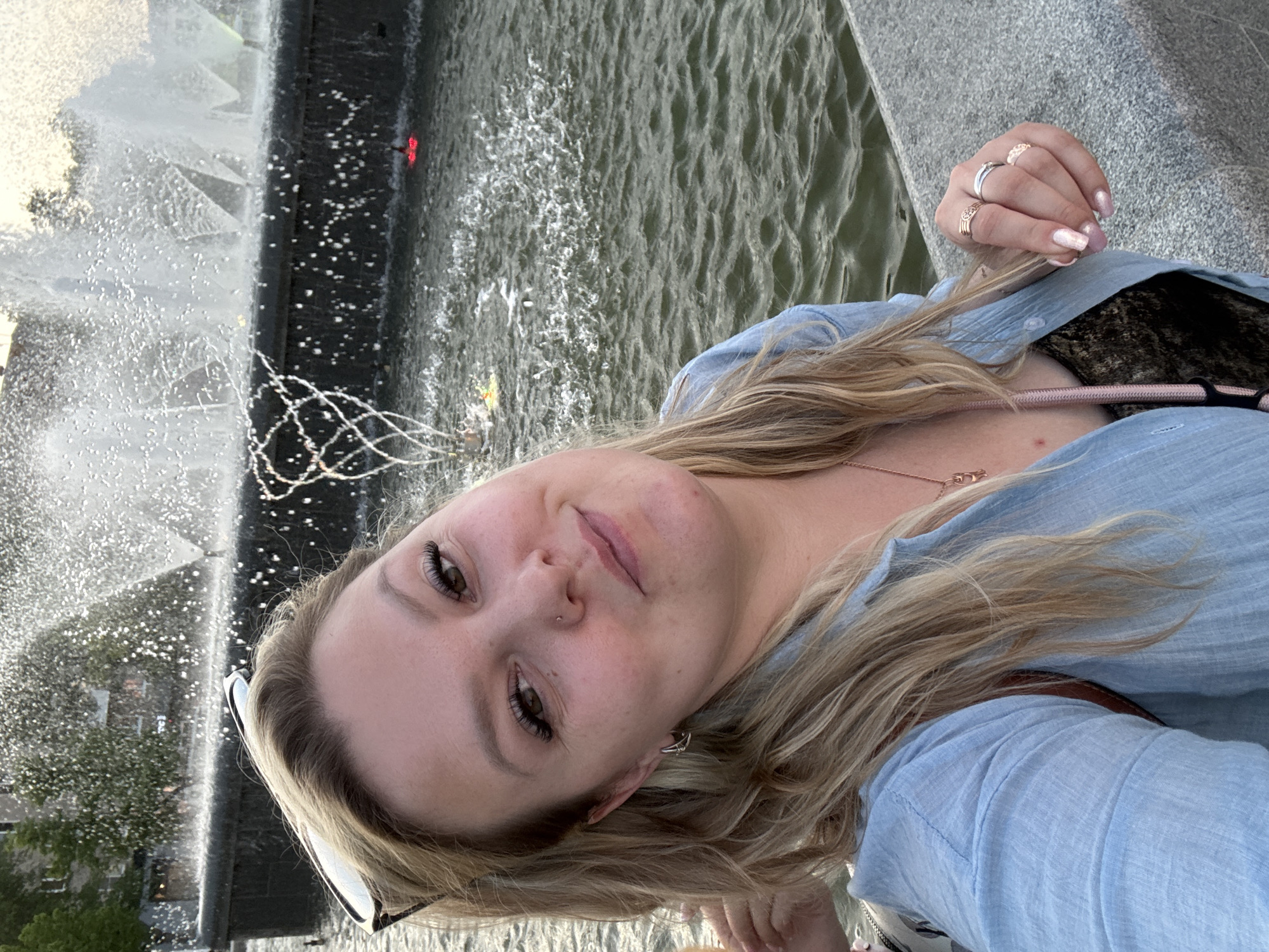A woman with blonde hair, wearing a light blue shirt, poses for a selfie. She stands next to a large fountain. Rings adorn her fingers. The background features a water feature with spouts and a pool. The location is Chaikovsky, Russia.