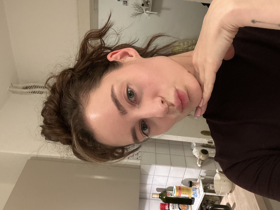 The image depicts a young adult female taking a selfie in a domestic kitchen environment within a residence in the city of Unknown, Unknown. The female, centered in the frame, has light skin, green eyes, and brown hair styled in a messy bun. She is wearing a dark, possibly black or dark brown, long-sleeved top. Her head is slightly tilted, and her lips are pursed in a "kissing" gesture. Her right hand is positioned under her chin, with her fingers extended and her palm resting against her jawline. Her fingernails are painted a light pink. A small, faint tattoo resembling a dotted line with an arrow at the top is visible on her left forearm/wrist area, near the bottom edge of the frame.
The background shows a kitchen area with white wall tiles and light-colored upper cabinets. On a countertop to the left, several items are visible, including a dark bottle of olive oil, a red-capped bottle of "Ramm" brand sauce, two cylindrical containers (one appearing to be salt), and a white mug with a dark rim. Below the countertop, a white pipe and a radiator-like fixture are partially visible against the tiled wall. Further back and to the right, a doorframe is present, and a shelf with decorative items, including what appears to be dried branches, is mounted on the wall. The lighting appears to be artificial and diffused, indicating an indoor setting. There are no other individuals or active interactions visible in the scene.