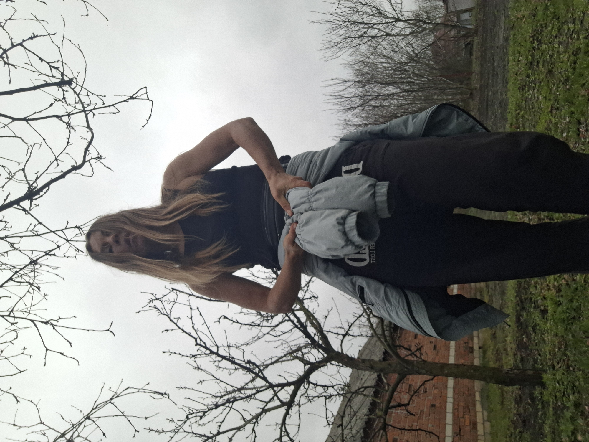 A woman with long blonde hair stands outdoors in Khlevnoye, Russia. She is wearing a black tank top and black pants and holding a light blue jacket. Bare tree branches frame the top and bottom of the image. In the background, there is green grass and a brick building. The sky is overcast.