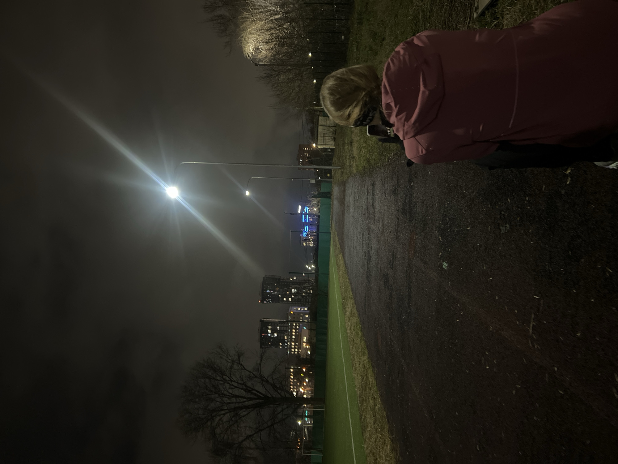 The image depicts an urban scene at night, likely in Kazan, Russia, given the architectural style visible in the background. A person wearing a pink coat is partially visible in the upper right. The environment features a dark, possibly wet path next to a green artificial turf or field. The cityscape in the distance is illuminated with various lights, indicating the presence of buildings and possibly street lighting. A bright streetlight is positioned to the left, casting a strong glare. A leafless tree is seen at the lower left. The sky is dark and cloudy. The person appears to be looking at something.