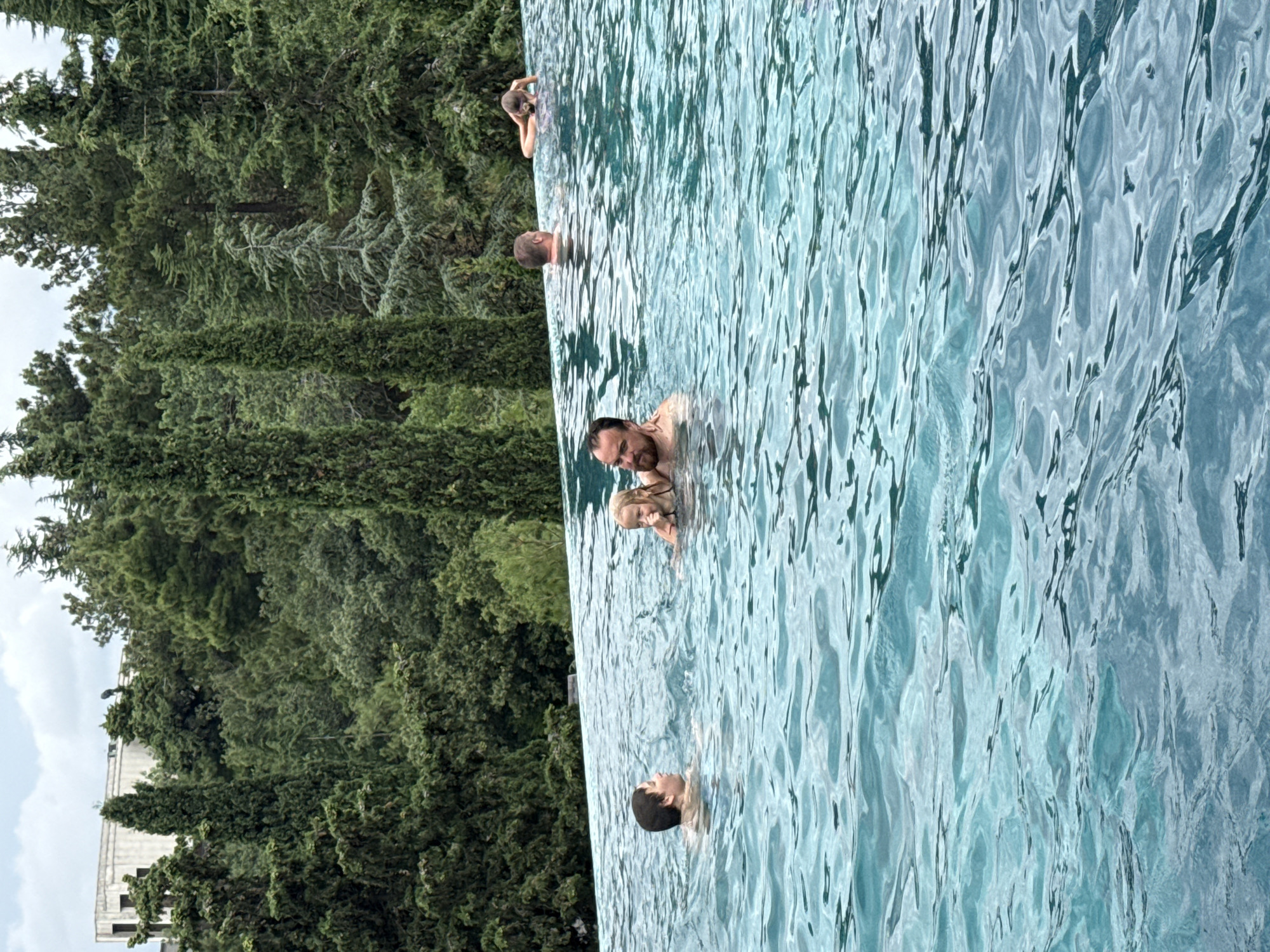 An outdoor infinity pool is filled with clear blue water. In the pool, five people are visible. In the foreground, a young boy is looking to his left, while a man with a beard is facing the viewer. Further back, a woman holds a young girl, who is looking at the camera. To the right, two men are partially submerged in the water. The pool is surrounded by a lush green forest, with tall cypress trees dominating the landscape. In the background, a modern, multi-story building with a light-colored facade is visible among the trees. The sky above is overcast with clouds.