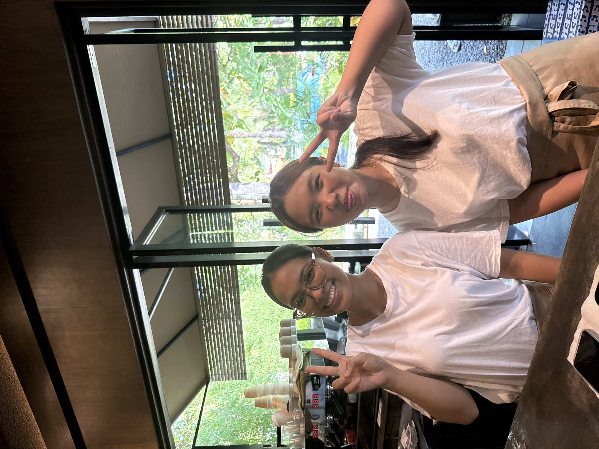 Two young women are captured in a vertical photo, both smiling broadly and making a peace sign (V-sign) with one hand. They appear to be posing behind a counter, likely in a cafe or similar establishment.
The woman on the left side of the frame (when viewing the image in its original, rotated state) has long, dark hair, wears a white t-shirt, and has a light-colored apron or skirt. Her left hand is raised, forming the peace sign. The woman on the right side of the frame wears glasses, has her dark hair pulled back, and is also wearing a white t-shirt. Her right hand is raised in a peace sign. Both women look directly at the camera with cheerful expressions.
In front of them, on the counter, a coffee machine with multiple buttons and nozzles is visible, along with stacks of disposable cups (some white, some brown). The background features large windows framed by dark metal, offering a view of lush green trees and foliage, suggesting a vibrant outdoor tropical setting. Vertical slatted blinds or bamboo screens are visible outside the windows, partially obscuring the greenery. The interior features dark wooden paneling on the walls.
The bright natural light indicates it is daytime. The women's poses and smiles suggest a friendly, casual moment, possibly taken during their workday. The presence of the coffee machine and cups strongly implies the setting is a cafe or coffee shop. No discernible text is visible in the image. The scene takes place in Kuta Selatan, Indonesia.
