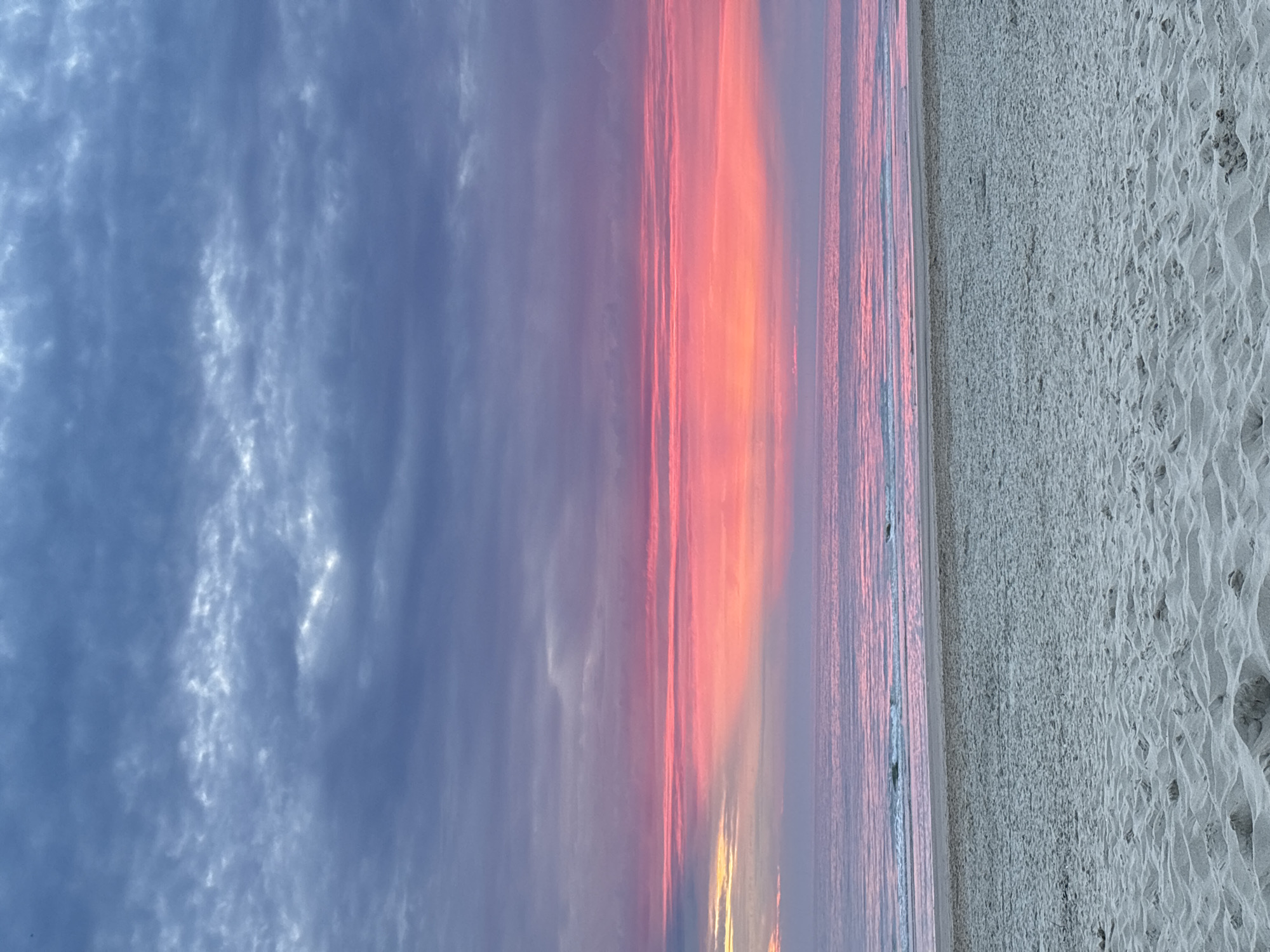 The image shows a beach at sunset. The sand in the foreground is light-colored and textured. The ocean is calm with small waves, reflecting the colors of the sky. The sky is filled with dramatic clouds in shades of purple, pink, and orange. The scene is peaceful and serene. This image could have been taken at the beach in Castricum, Netherlands, which is known for its beautiful sunsets and long stretches of sand.