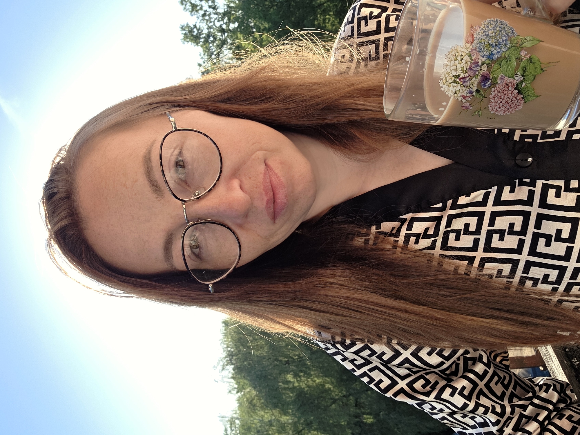 A woman with long, auburn hair and round glasses holds a glass of coffee or tea with a floral design. She wears a black and white patterned shirt. The background shows trees and a blue sky, suggesting an outdoor setting. While the exact location in Chaikovsky, Russia is not evident, the presence of green vegetation and a clear sky point to a possibly suburban or park-like environment.