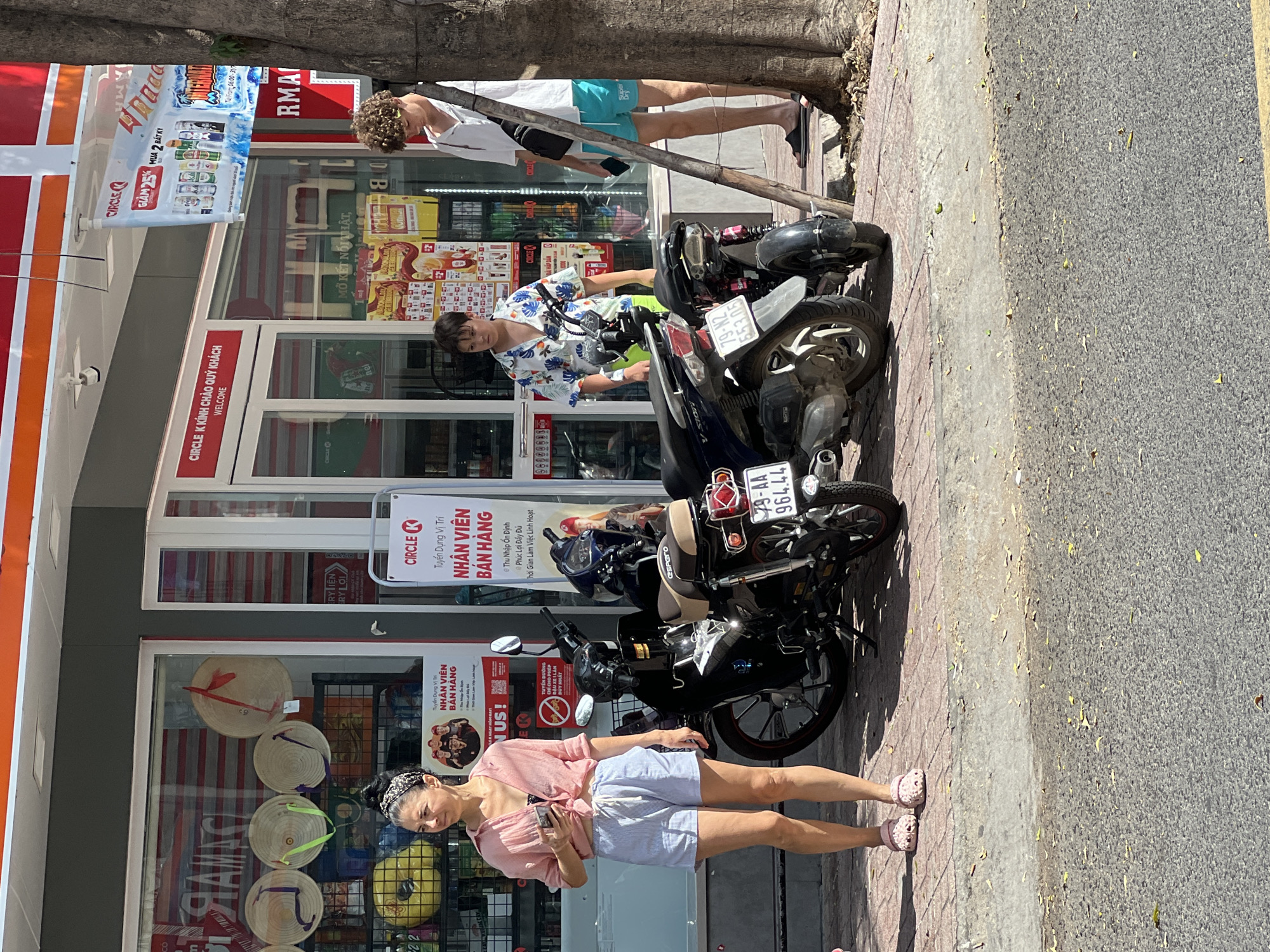 The image depicts a sunny outdoor scene in front of a Circle K convenience store in Nha Trang, Vietnam. Three people are visible. On the left, a woman with dark hair pulled back in a bandana, wearing a light pink unbuttoned shirt over a dark top and light blue striped shorts, stands holding a mobile phone. To her right, a younger person with dark hair wears a short-sleeved patterned shirt with a floral design and light green shorts. Further to the right, a young man with curly brown hair is dressed in a white short-sleeved shirt and turquoise swim trunks. Two motorcycles are parked on the sidewalk. The Circle K store has large glass windows displaying various products and signage, including Vietnamese text indicating recruitment and promotions. The ground is paved with red brick and concrete, with fallen leaves scattered about. A large tree trunk is visible on the right side of the frame. The overall atmosphere is casual and appears to be a typical day in a Vietnamese city.