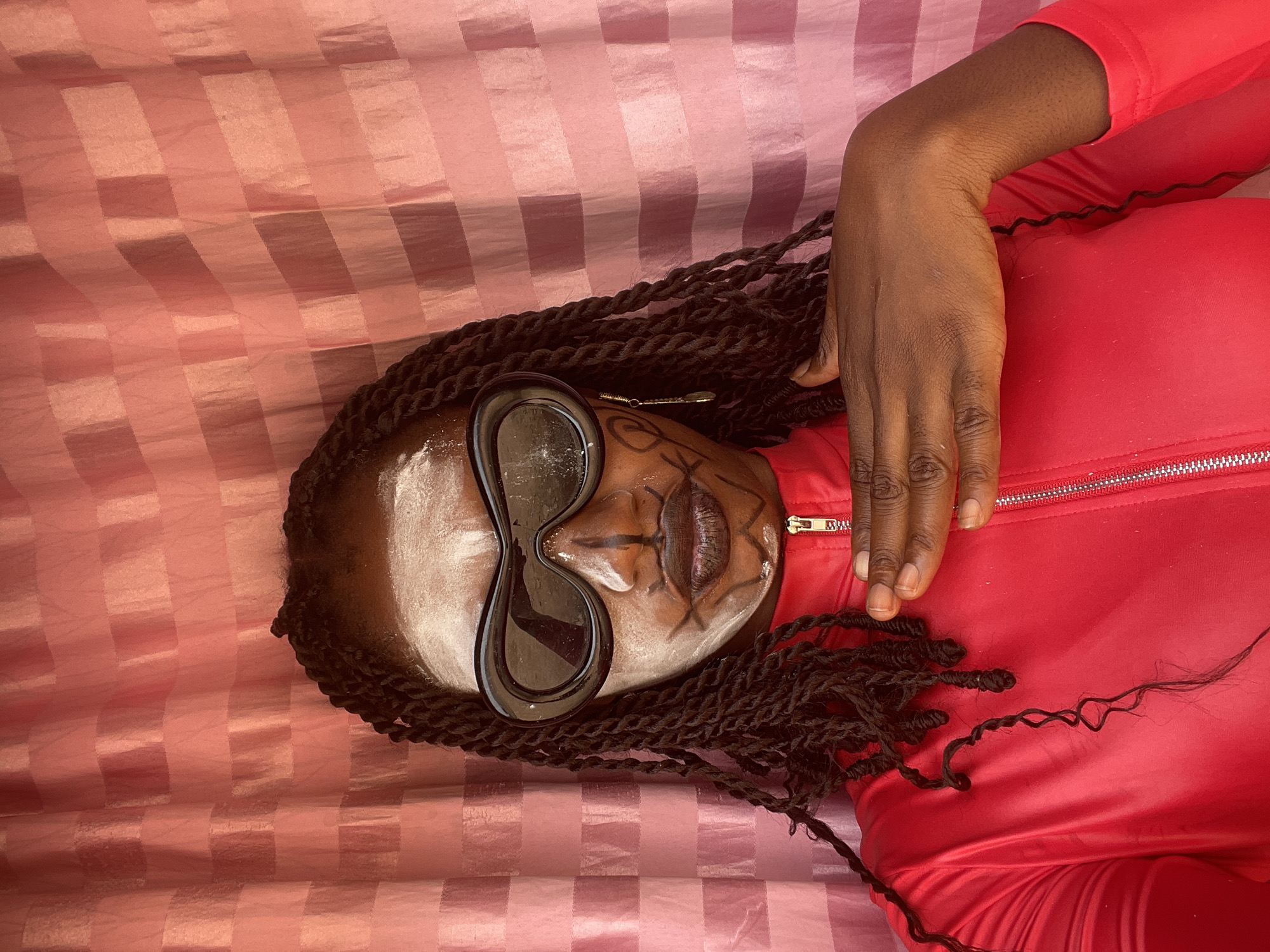 A person is depicted lying on their back, with their head positioned towards the left side of the frame and their body extending to the right. The individual has dark skin and long, dark braided hair that is spread around their head and shoulders. They are wearing large, dark, uniquely shaped sunglasses that cover their eyes.
Distinctive face paint or makeup is visible on their face. A white, powdery substance is applied to the forehead, nose, and upper cheeks, contrasting with their skin tone. Dark lines and intricate patterns are drawn on the face, including around the mouth and extending onto the cheeks. The lips are painted a dark, possibly reddish-brown or black color. A small, metallic hair accessory can be seen among the braids near the person's right temple.
The person is dressed in a bright red long-sleeved top, likely a jacket or tracksuit, featuring a prominent metallic zipper down the front. A dark-skinned hand and forearm are visible in the upper right portion of the image, resting on the red clothing.
The individual is resting on a surface covered with a light pink fabric. This fabric has a noticeable checkered or striped pattern created by alternating squares or rectangles of different textures, possibly matte and glossy, giving it a subtle three-dimensional effect.
The scene appears to be a posed photograph, with the person lying still. The overall aesthetic suggests an artistic or stylized portrait. The setting is in Akwanga, Nigeria. There is no visible text in the image.