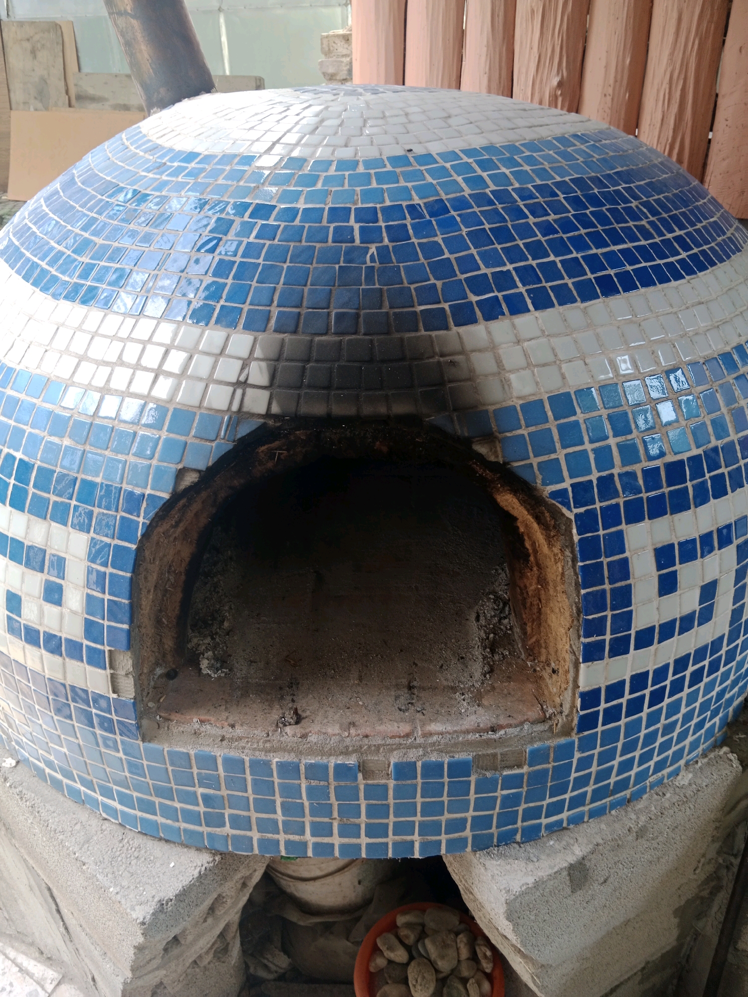 A dome-shaped tiled oven with blue and white tiles is positioned outdoors. The oven has a circular opening in the front. The oven is placed on concrete blocks. In the bottom right corner of the image, a small orange bowl filled with pebbles is visible. The background shows wooden structures and some discarded materials.
