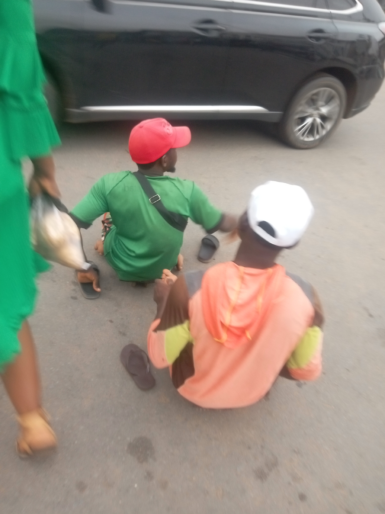 The image depicts two men sitting on the ground in Fegge, Nigeria. One man is wearing a green shirt, a red cap, and a black shoulder bag. The other man is wearing a peach-colored hoodie and a white cap. Both men have their sandals off. A black car is parked in the background. A person wearing a green dress and carrying a plastic bag is walking past. The ground is asphalt.