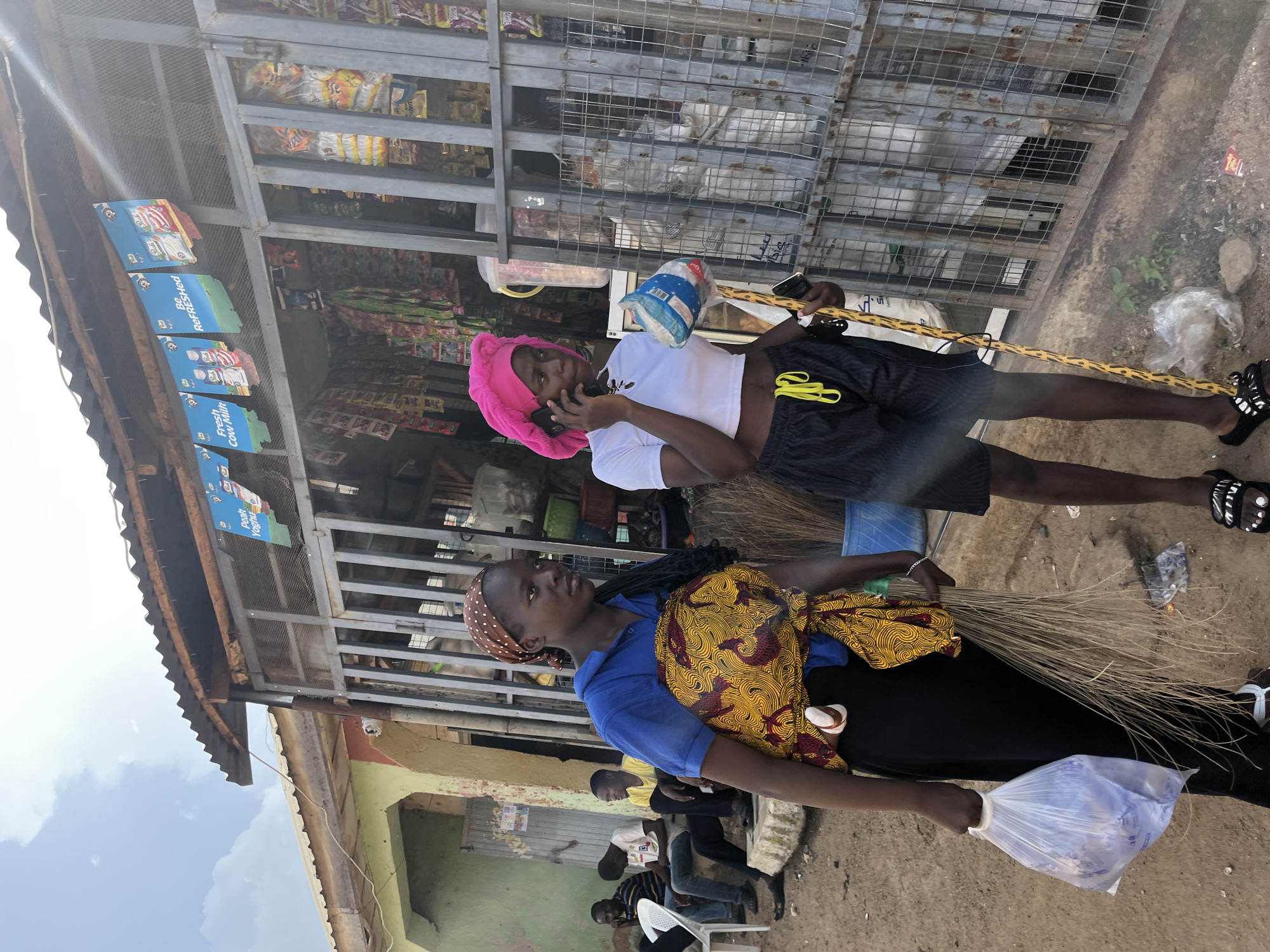 In Jos, Nigeria, on a dirt road, a street vendor's stall is depicted. The stall has a corrugated metal roof supported by wooden beams. Behind a metal grate, shelves are stocked with various goods, including bags of snacks and drinks. Two young women are the primary subjects in the foreground.
The woman on the left, facing left, wears a blue t-shirt and a brown headscarf. She carries a large plastic bag filled with items in her left hand and a bundle of brooms in her right. A brightly colored yellow and red fabric, likely a wrap, is tied around her waist, carrying a baby.
The woman on the right, facing left, is wearing a white crop top, black shorts with yellow drawstrings, and a pink headscarf. She is holding a white container in her left hand and a mobile phone to her ear in her right. A yellow and black patterned stick is held vertically in her right hand, near the phone. She is speaking on the phone and looking towards the stall.
In the background, to the left of the women, several men are seated on a low concrete structure. They appear to be customers or passersby. The environment is dusty, with the ground composed of dirt and small stones. Various discarded plastic bags and wrappers are visible on the ground. The overall scene captures a slice of daily life at a marketplace in Jos.