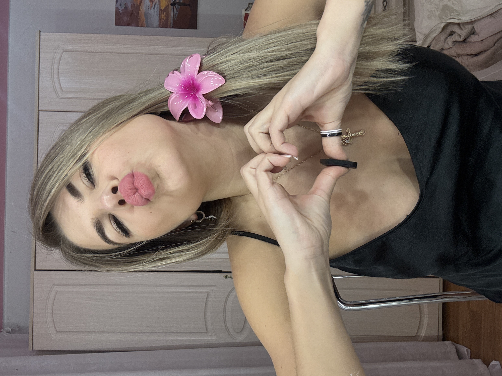 A female individual is depicted in a close-up, chest-up indoor portrait, identified as being in Chaikovsky, Russia. She has light skin, long blonde hair with highlights, and is wearing a black tank top or slip dress with thin straps. Her eyes are closed, and her lips are pursed in a kiss gesture, enhanced with pink lipstick. A bright pink floral hair clip is secured on the left side of her head. Her hands are positioned at chest level, with thumbs and index fingers forming a heart shape. A thin gold-colored chain with a small cross pendant hangs around her neck. On her right ring finger, she wears a black ring and a silver-toned ring with a black band. On her left ring finger, a black elastic band is present. Her fingernails are painted white. A metal chair back is partially visible on her left side, indicating she is seated. The background features light-colored wooden wardrobe doors with recessed panels, a glimpse of a pink wall section, and a framed artwork depicting a sunset over water with a silhouetted figure on the far right. A light-colored curtain is visible on the far left, and a bed with light-colored bedding is partially visible in the lower right.