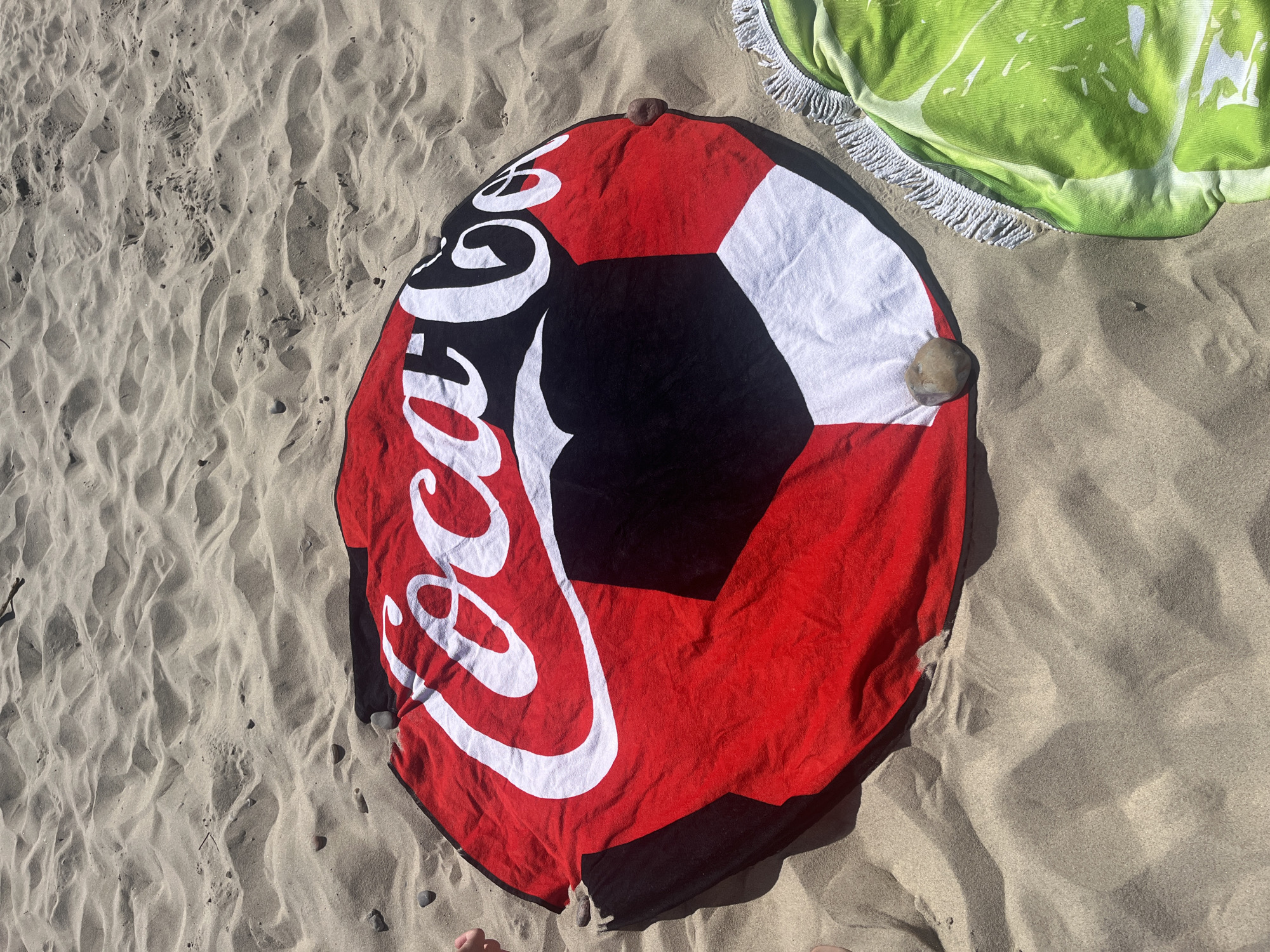 This media file shows a top-down view of two round beach towels on a sandy beach. The primary subject is a circular beach towel with a Coca-Cola logo in white script against a black and red background, designed to resemble a soccer ball. The towel is partially folded and has a few small stones holding it down. To the upper right of the Coca-Cola towel is another round towel, primarily bright green with a white pattern resembling lime slices and a white fringe border. The towels are spread out on a sandy surface that shows the texture of footprints and ripples, indicating it's a beach. The lighting suggests it is daytime, likely a sunny day given the shadows cast. The scene implies a leisure activity, such as sunbathing or relaxing at the beach. No people are clearly visible, though parts of feet are visible at the bottom edge of the frame. The visible text is the "Coca-Cola" logo. The setting is a beach, with the sand being the dominant ground element. While the prompt specified Zelenogradsk, Russia as a location context, there are no specific visual cues in the image to confirm this location.
