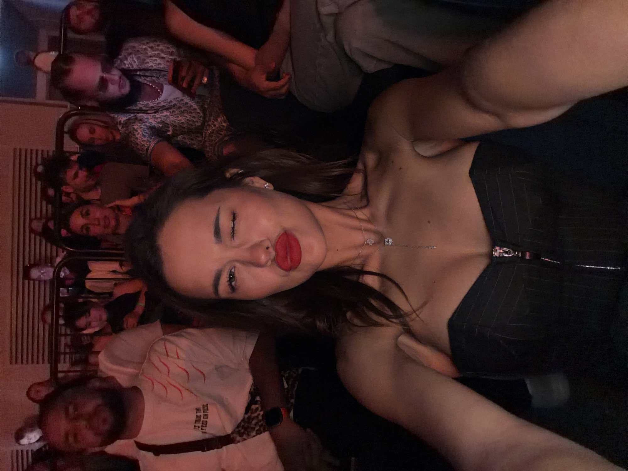 The image depicts an indoor social gathering or entertainment event, potentially a concert or club, located in Khimki, Russia. The lighting is dim with a prevalent red and orange ambient hue.
In the foreground, a young adult female with long dark hair is captured in a selfie-style photograph. She is winking with her right eye and puckering her lips, which are coated in bright red lipstick. She wears a strapless, dark pinstriped top with a metallic zipper down the front, and a delicate silver necklace featuring multiple pendants, including a square and a clover design. Her left arm, extended to hold the camera, is visible.
Behind the primary subject, in the midground, several individuals are seated or standing. To the left, a male with a beard and dark hair wears a white t-shirt with red wavy lines and partial text reading "IS TRUE THE...FEED ON MUSL." He has a dark crossbody bag with a leopard print strap across his chest and an orange wristband or watch on his left wrist. He is observing the general direction of the event.
To the right of the primary subject, another male, characterized by dreadlocks or braids and dark sunglasses, is engrossed in a smartphone with a prominent orange case. He wears a patterned shirt and a heavy chain necklace, with a ring visible on his right hand. Adjacent to him, a woman with blonde hair looks forward. Further to the left and slightly behind, a woman with dark hair and a bright pink top is partially visible.
In the background, additional individuals are visible in tiered seating behind a dark metal railing, suggesting an upper level or balcony. Their features are less distinct due to distance and low light, but they appear to be observing the event or engaged with personal devices. The immediate background also shows textured wall panels.