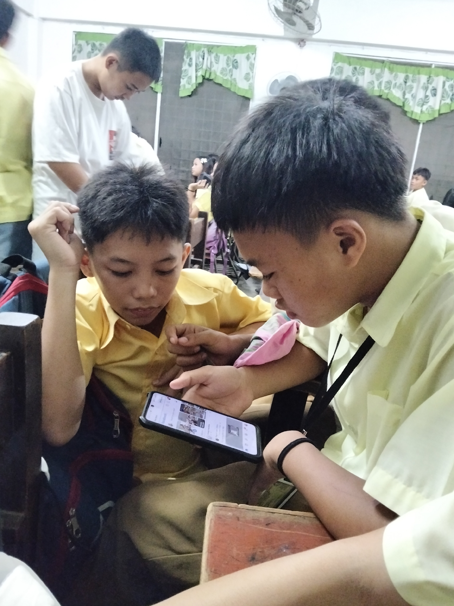 Two young Filipino boys, likely students in Davao City, are engrossed in a smartphone. One boy, wearing a light yellow collared shirt, is holding the phone and showing its screen to the other boy, who is also in a similar yellow shirt and looking down at the device. Their focused expressions suggest they are engaged in a game or browsing content. The background hints at a classroom or common area, with a ceiling fan visible and green and white curtains adorning a window. Other students and possibly an adult are faintly visible in the background, suggesting a school environment. The scene appears to be taking place during daylight hours, given the general lighting. The mood is one of shared concentration and perhaps a break from school activities. There is no visible text in the image.
