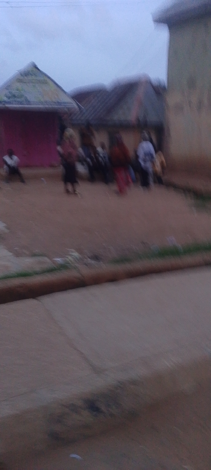 The image depicts an outdoor scene in what is identified as Gura Top, Nigeria. The setting appears to be a village or informal settlement with modest structures and unpaved ground. Several individuals are visible, though their features are indistinct due to motion blur and the overall low resolution of the photograph.
In the background, there are buildings with pitched roofs. One structure to the left has a prominent triangular roof and a pink facade. Adjacent to it, another building displays a darker, possibly corrugated, roof. To the right, a building with a lighter, textured wall is partially visible, suggesting it might be made of concrete or stucco.
The ground in the foreground and mid-ground is composed of dirt or sand, with some areas appearing damp or muddy, especially in the lower portion of the image. A thick, cylindrical object, possibly a log or a raised edge of pavement, runs horizontally across the mid-ground, separating the foreground from the area where the people are gathered.
The people in the scene are in motion, making it difficult to discern specific activities or interactions. However, their groupings suggest a social gathering or some form of community event. One individual on the far left appears to be seated or leaning. In the central area, a cluster of people are standing, with clothing in various colors, including red and white. The overall impression is of a moment captured during an ordinary day in this Nigerian location.