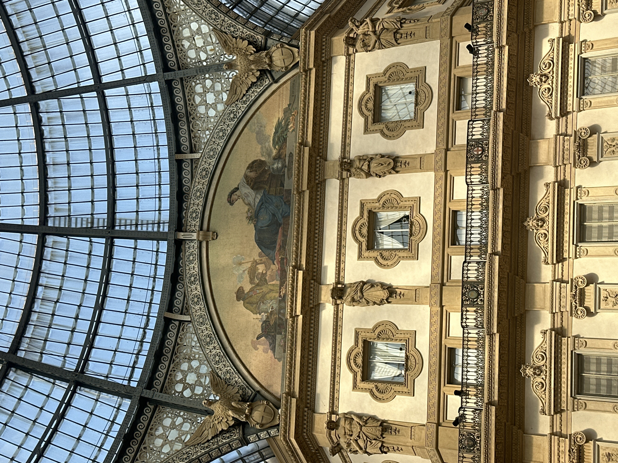 The image captures an interior view of the Galleria Vittorio Emanuele II in Milan, Italy, focusing on the ornate architecture and the grand glass-and-iron dome above. The scene is dominated by cream-colored walls adorned with classical sculptures and intricate decorative elements. A prominent semicircular mosaic depicts two figures in historical attire, with a man standing and a woman seated. Flanking the mosaic are golden eagle statues perched on pedestals, their wings spread wide. Below, a row of windows with patterned frames is visible, some with white curtains drawn. A black wrought-iron balcony with decorative panels and circular emblems runs across the middle of the structure. The uppermost section of the image shows the vast, segmented glass dome of the Galleria, allowing natural light to filter through. The overall impression is one of opulence and historical grandeur.