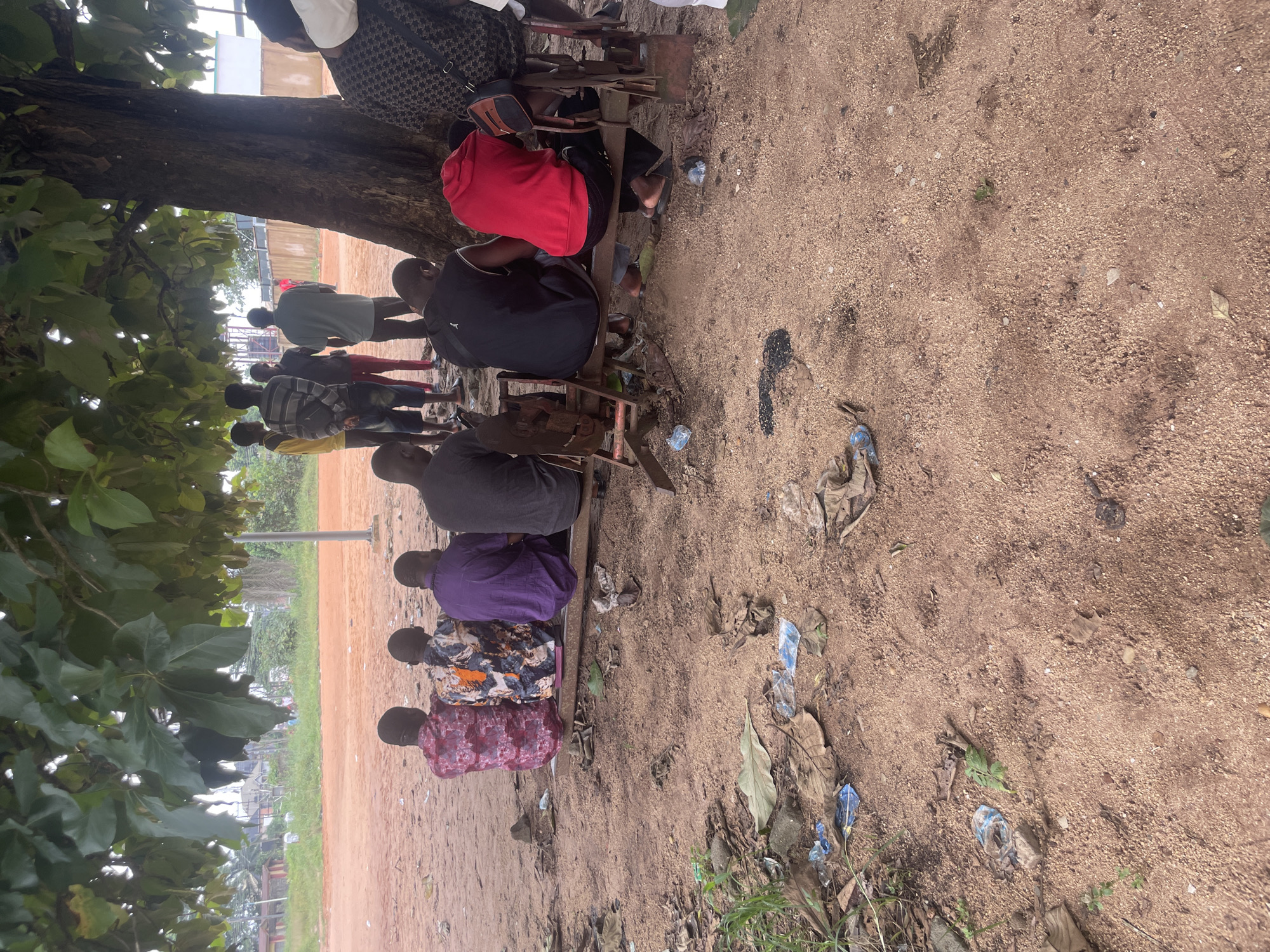 A group of people are sitting and standing under a large tree in what appears to be a public area in Ihiagwa, Nigeria. The ground is covered in reddish-brown dirt, with scattered leaves and plastic litter. Several individuals are seated on a low wooden bench, facing away from the camera. One person is wearing a vibrant red hooded sweatshirt, another a black sleeveless shirt, and others are in patterned shirts or darker clothing. Standing behind the seated group, a few individuals are visible, one in a yellow shirt and another in a striped shirt. In the background, there are some buildings and vegetation, suggesting an outdoor urban or semi-urban environment. The lighting indicates it is daytime, possibly overcast.