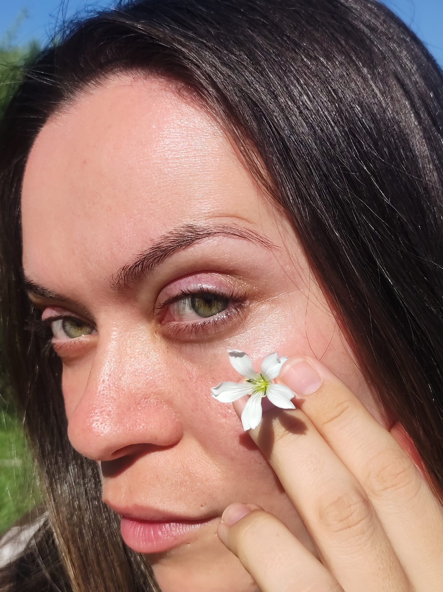 The image is a close-up, low-angle outdoor portrait of a person with dark brown hair and green eyes, holding a white flower with five petals and a green center to their cheek. The person is looking slightly to the left, with their right eye more prominent. The background consists of blurred green foliage and a clear blue sky, suggesting a sunny day. There are no identifiable landmarks or structures that would indicate a specific location within the city of Фоки, Russia. The lighting suggests the sun is to the left of the frame, casting a shadow from the flower onto the person's cheek. The person's skin appears smooth and slightly dewy.